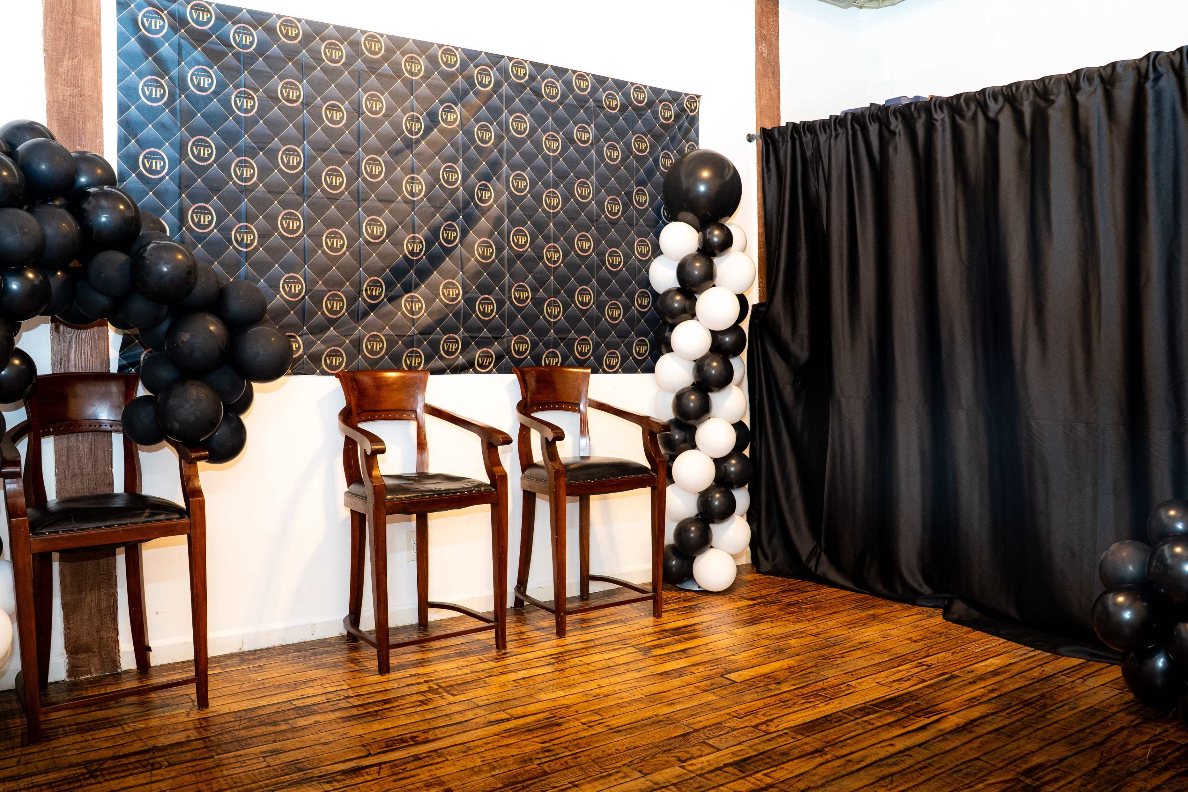 The image shows a decorated room with black and white balloons, two wooden chairs, and a black curtain, set against a patterned backdrop.