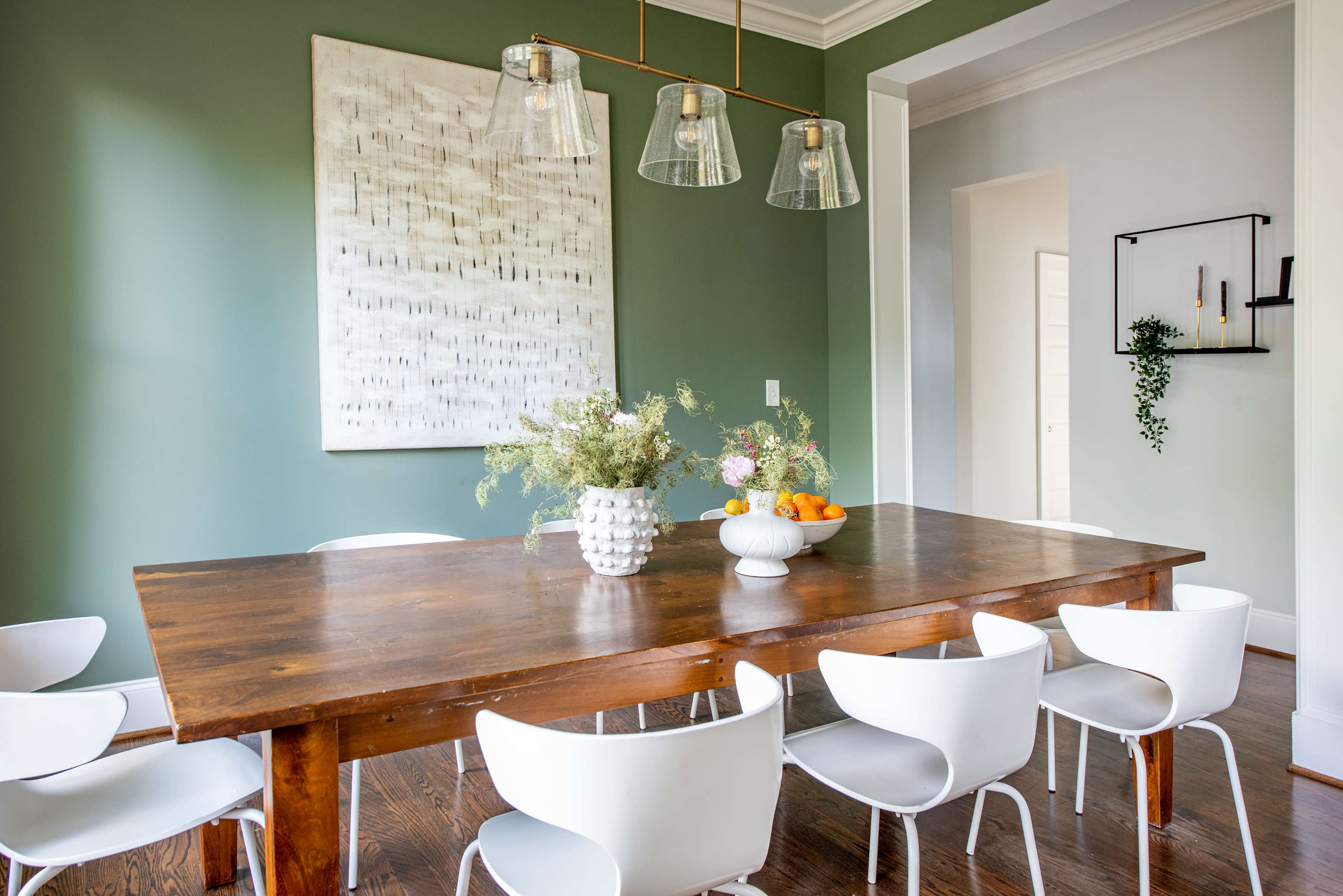 A wooden dining table is set with a vase of flowers and a bowl of fruit, surrounded by white chairs in a green-walled room featuring modern pendant lighting.
