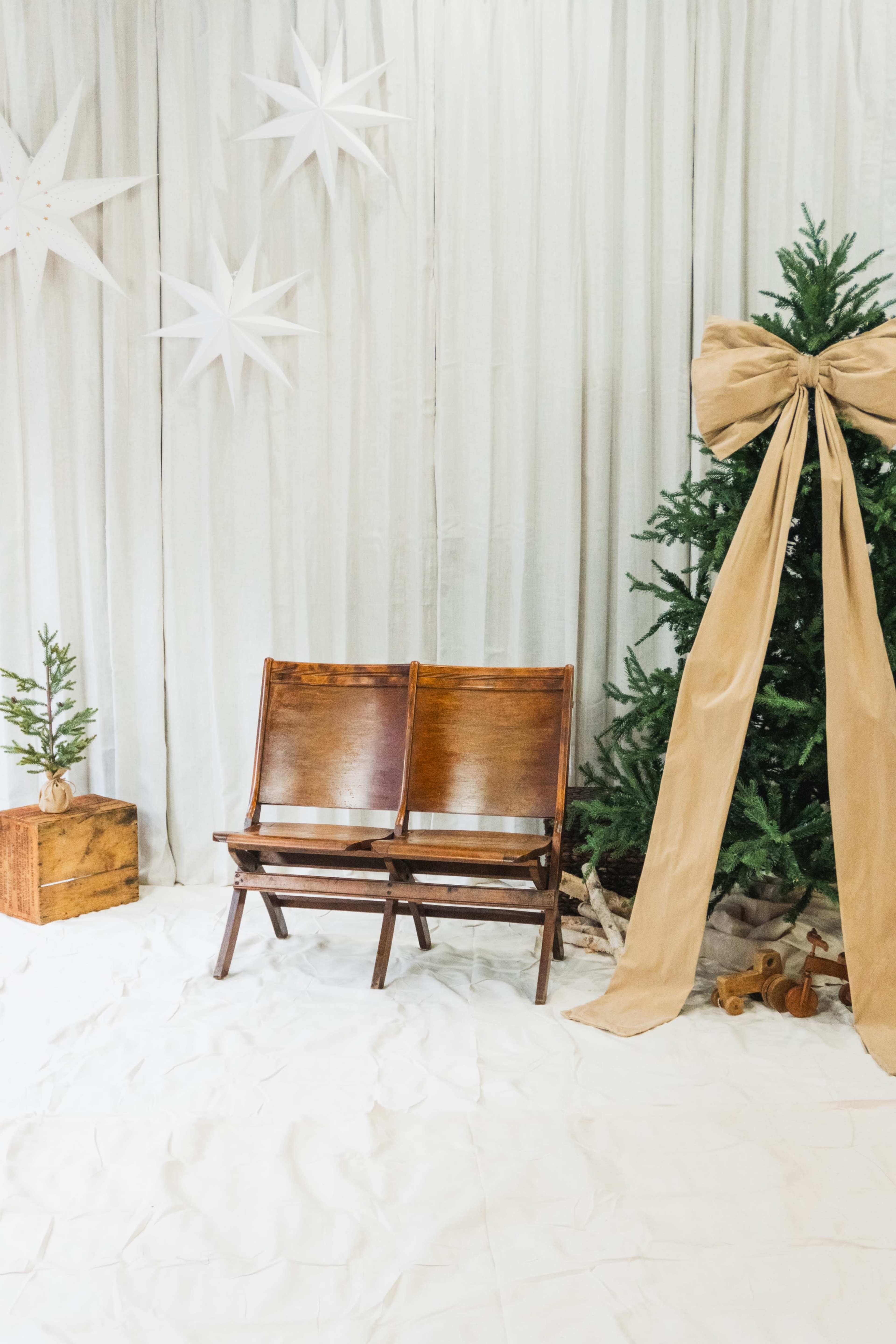 The image shows a simple indoor scene featuring two wooden chairs next to a Christmas tree adorned with a large bow and accompanied by minimalist decorations on a white backdrop.
