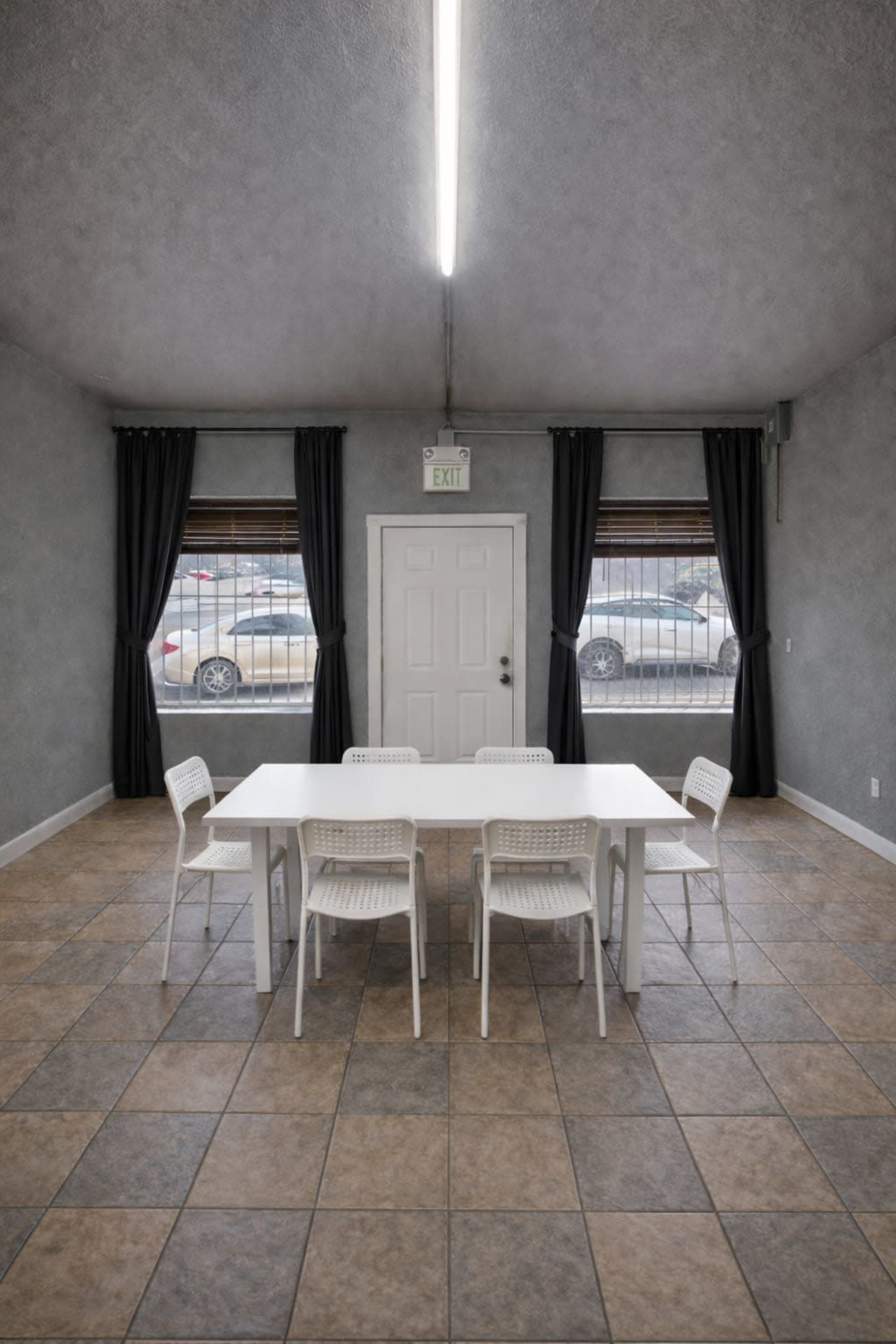 A simple room features a white table surrounded by four chairs, with two windows and a door visible in the background.