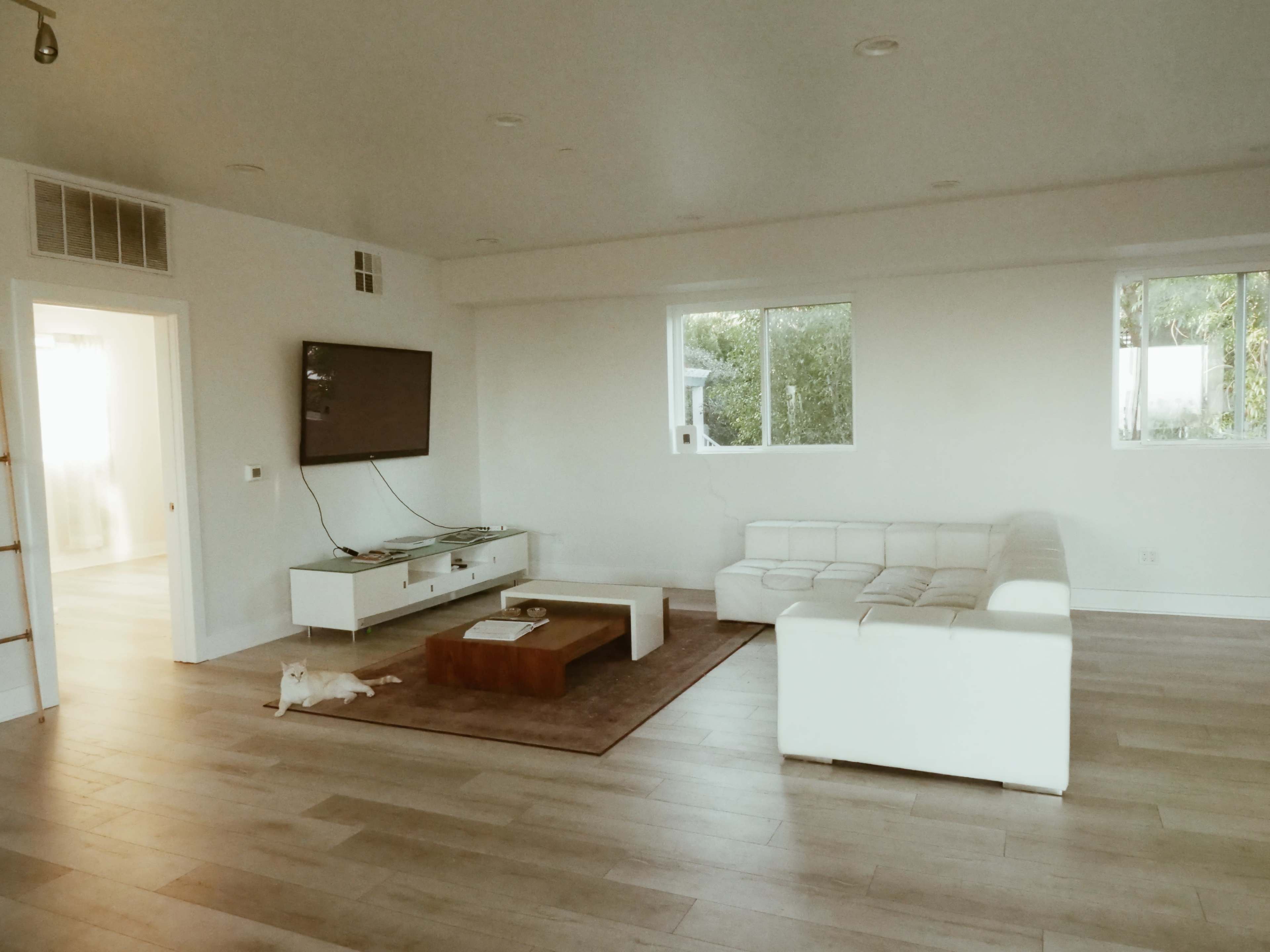A spacious living room features a white sectional sofa, a coffee table, a wall-mounted TV, and two large windows.