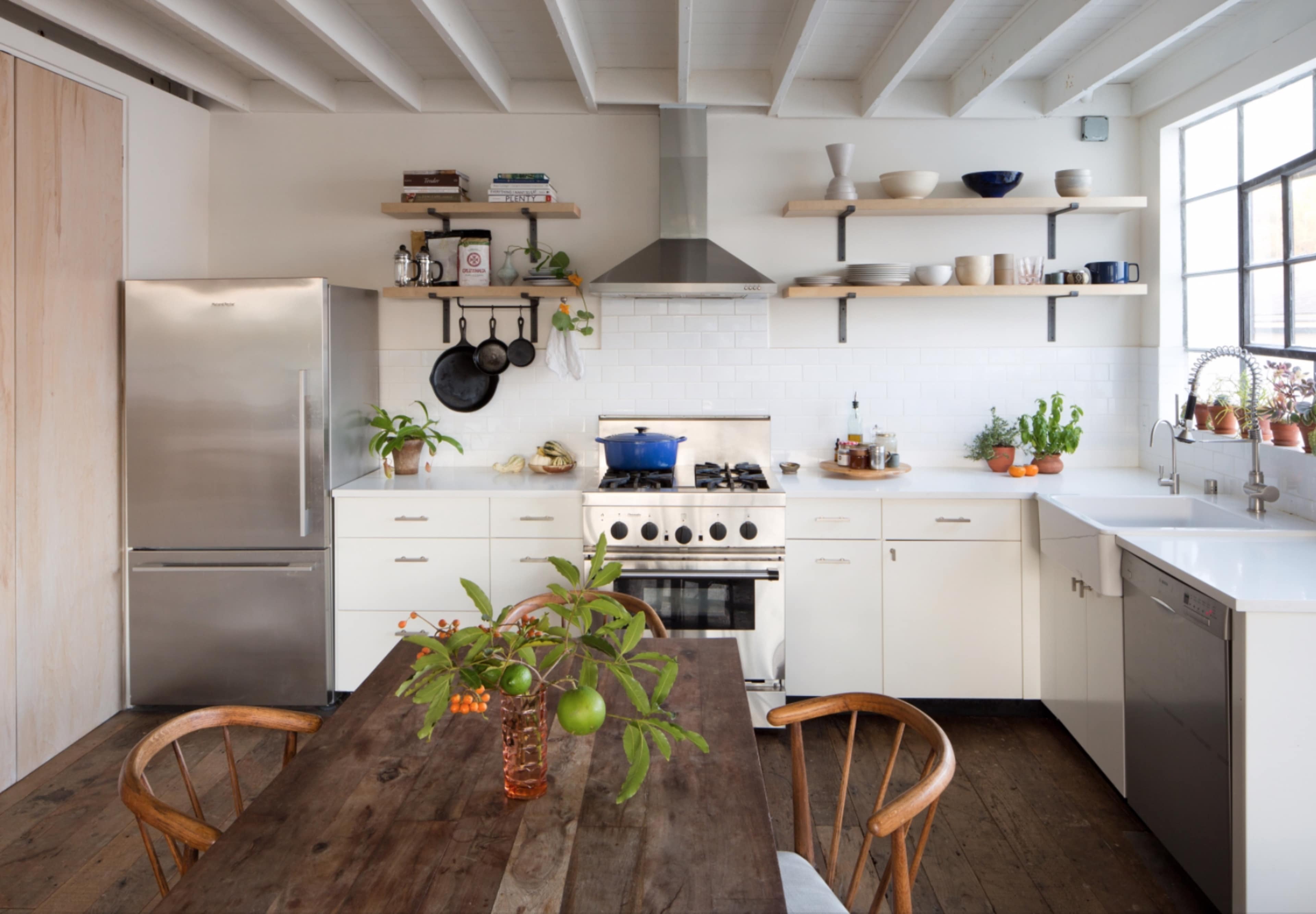 A modern kitchen features a stainless steel refrigerator, white cabinets, a gas stove with a hood, open shelving, and a wooden dining table adorned with a vase of plants.