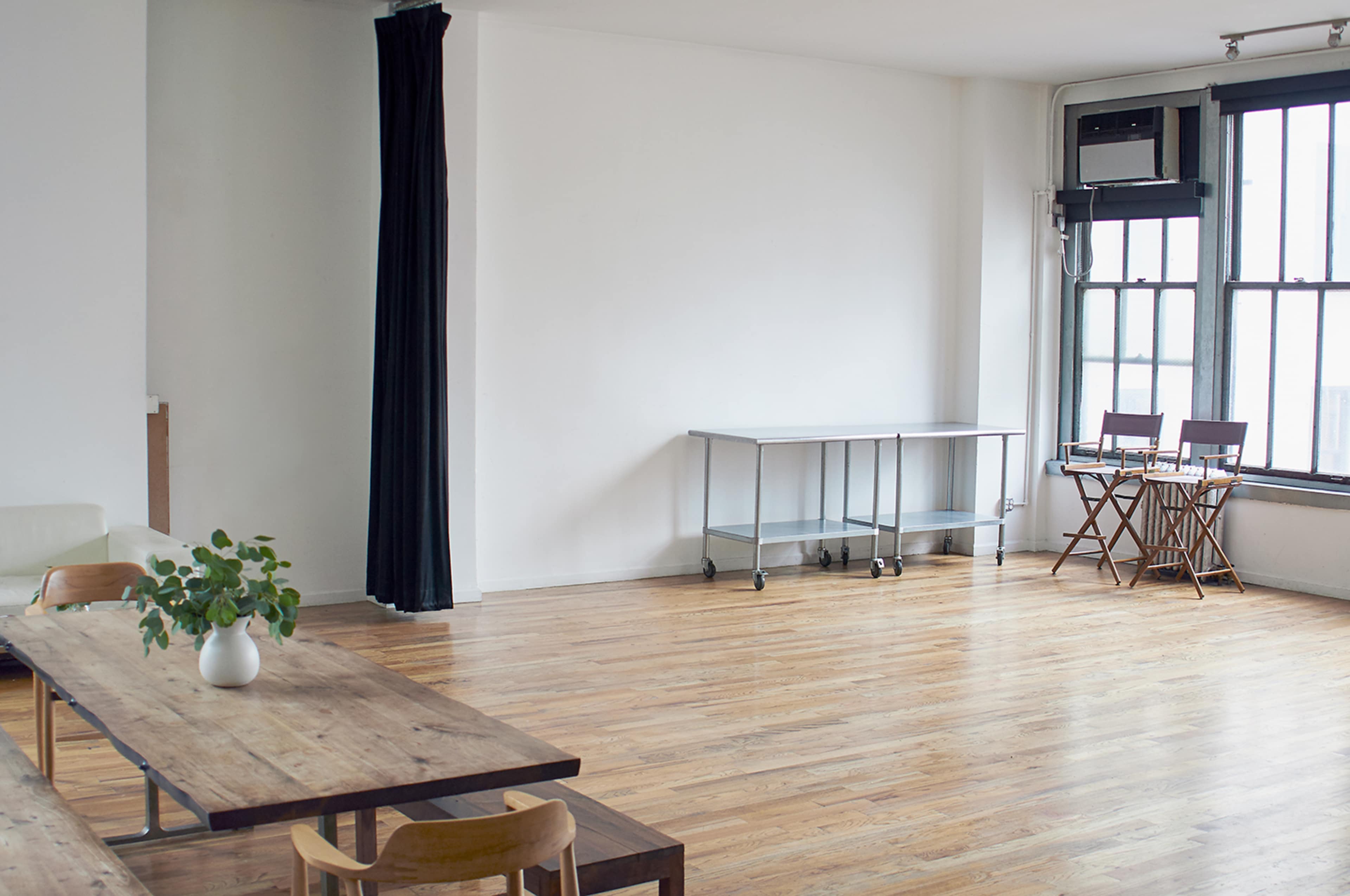 A minimalist room features wooden flooring, a large table, a steel cart, and two folding chairs near a window.