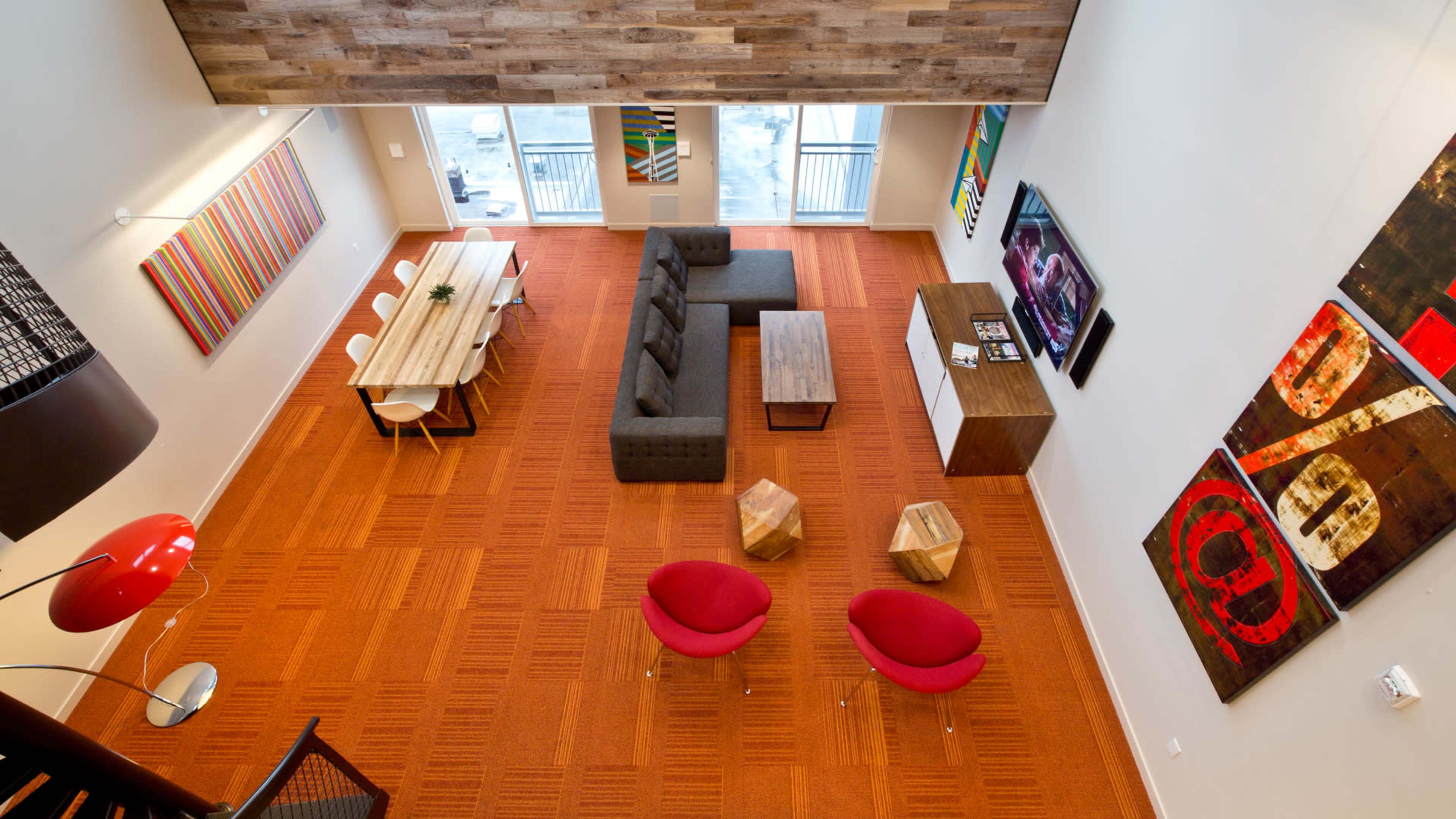 The image shows a modern living space featuring a spacious layout with a large sofa, a wooden dining table, red accent chairs, and colorful wall art.