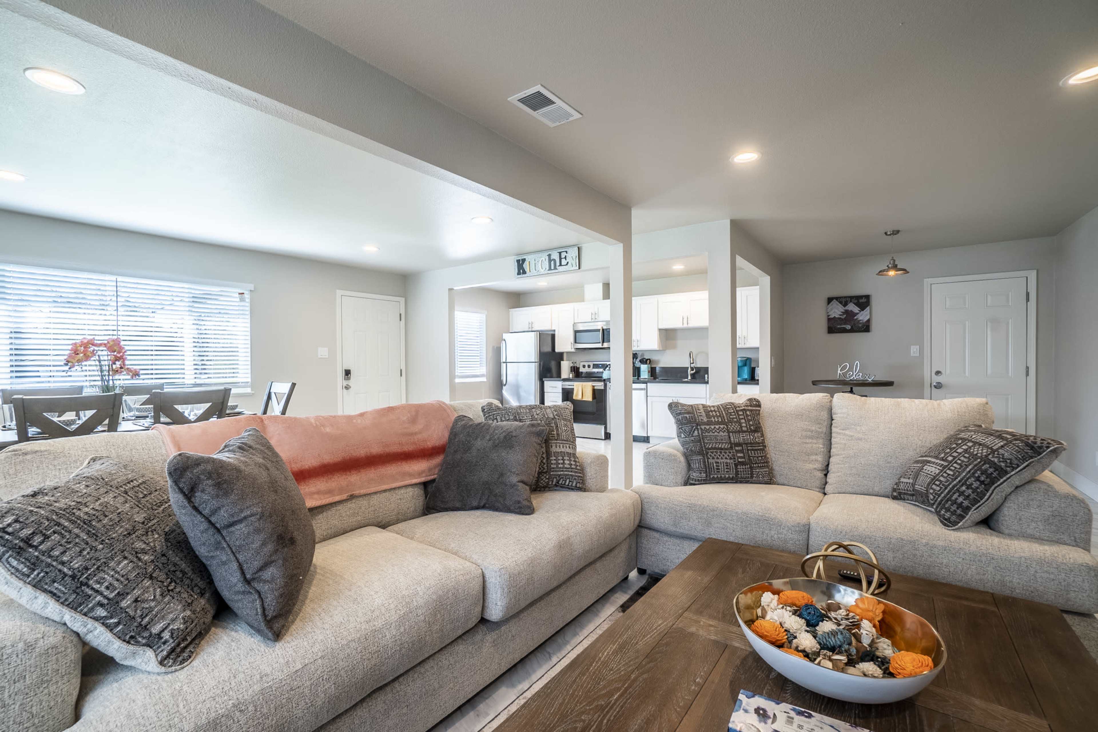 A modern living room with a light gray sofa, a wooden coffee table adorned with decorative items, and an open kitchen visible in the background.