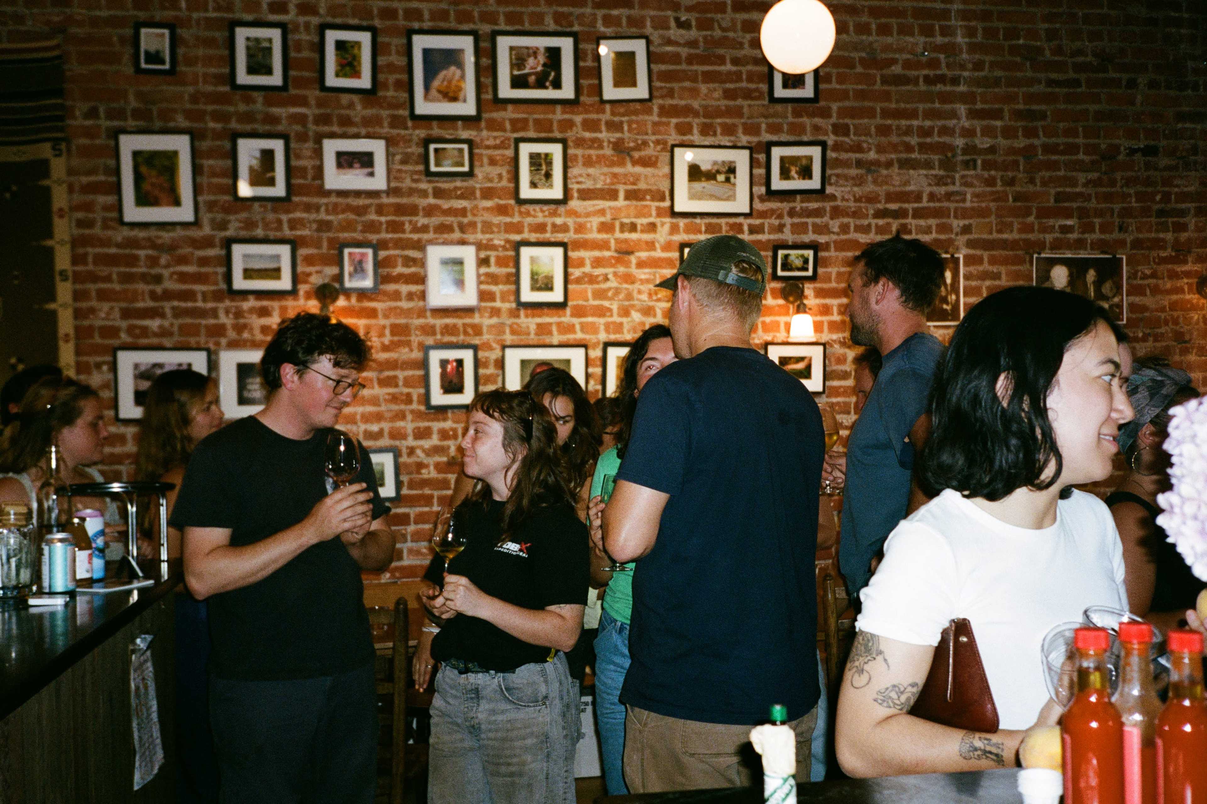 A group of people socializes inside a brick-walled bar, surrounded by framed photographs.