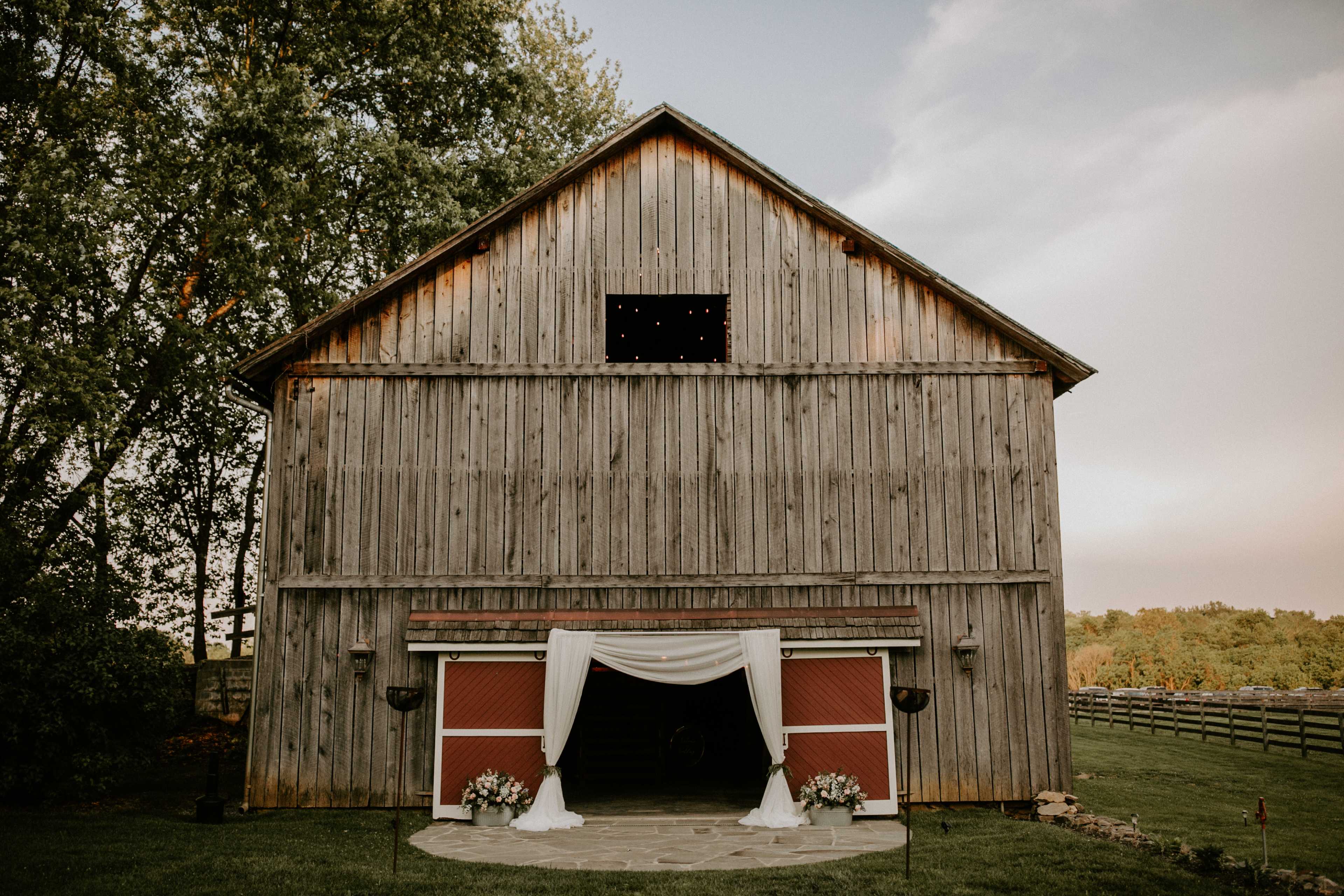 A rustic barn with a draped entrance and floral arrangements is set against a wooded landscape.