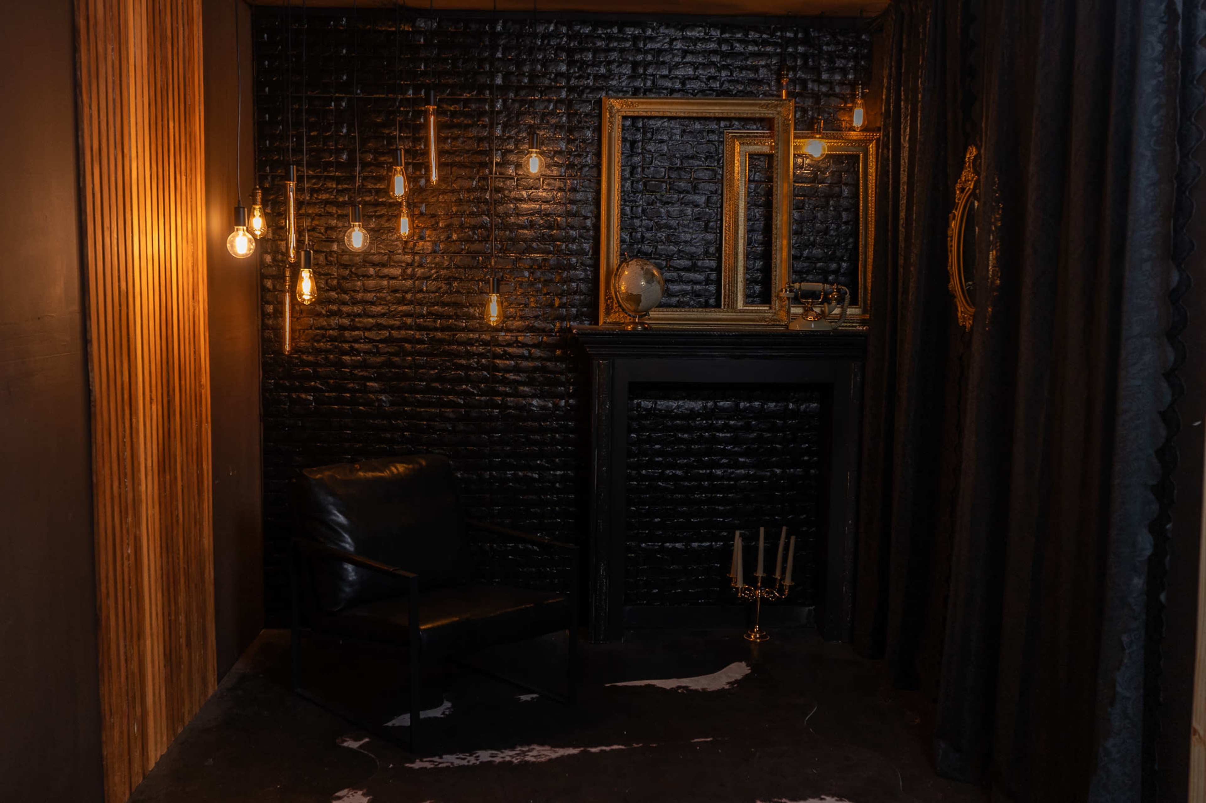 A dimly lit corner of a room featuring a black brick wall, a black armchair, hanging light bulbs, a decorative mirror, and a fireplace with a candle holder.
