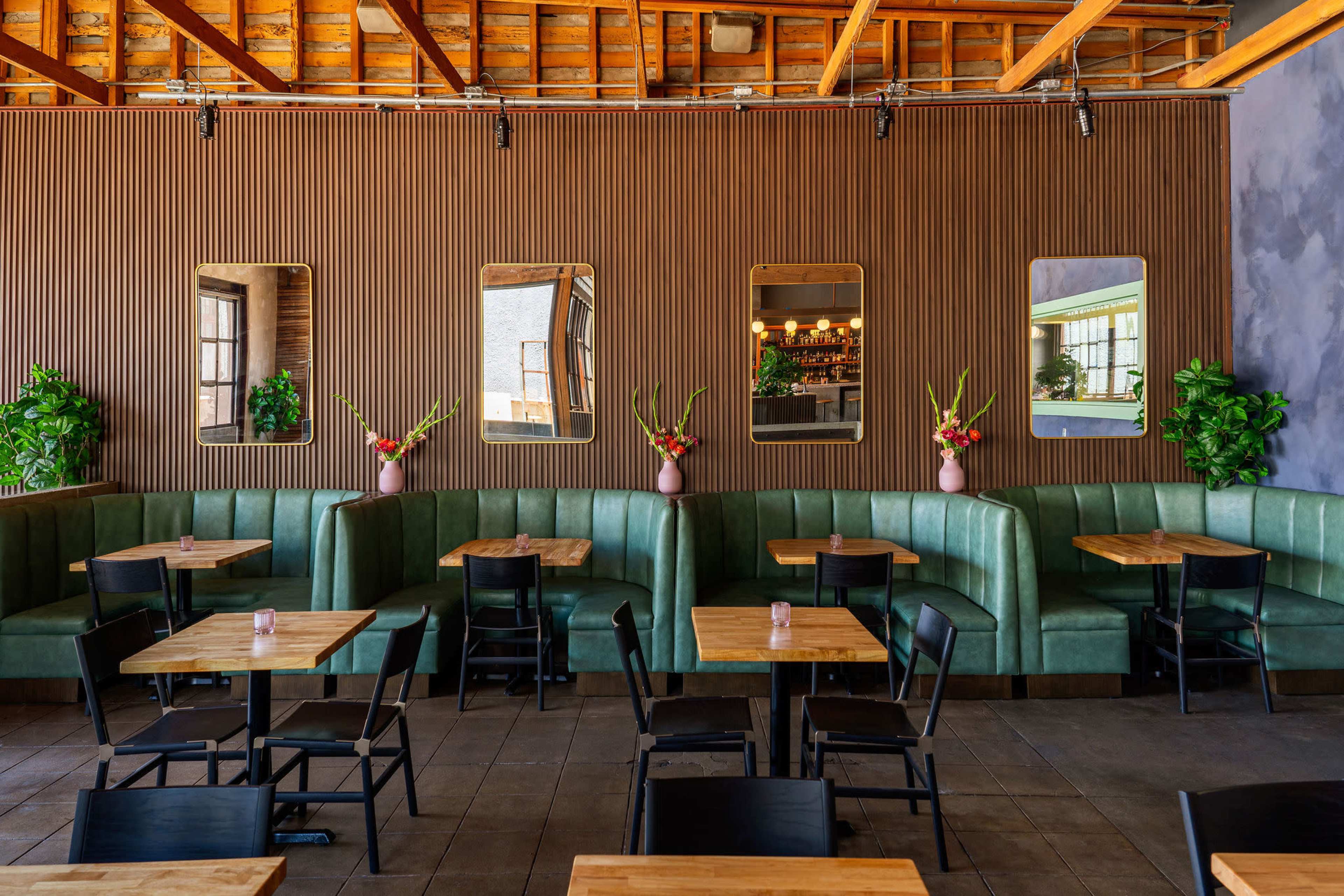 A modern restaurant interior with green upholstered booths, wooden tables, and decorative mirrors on the wall.