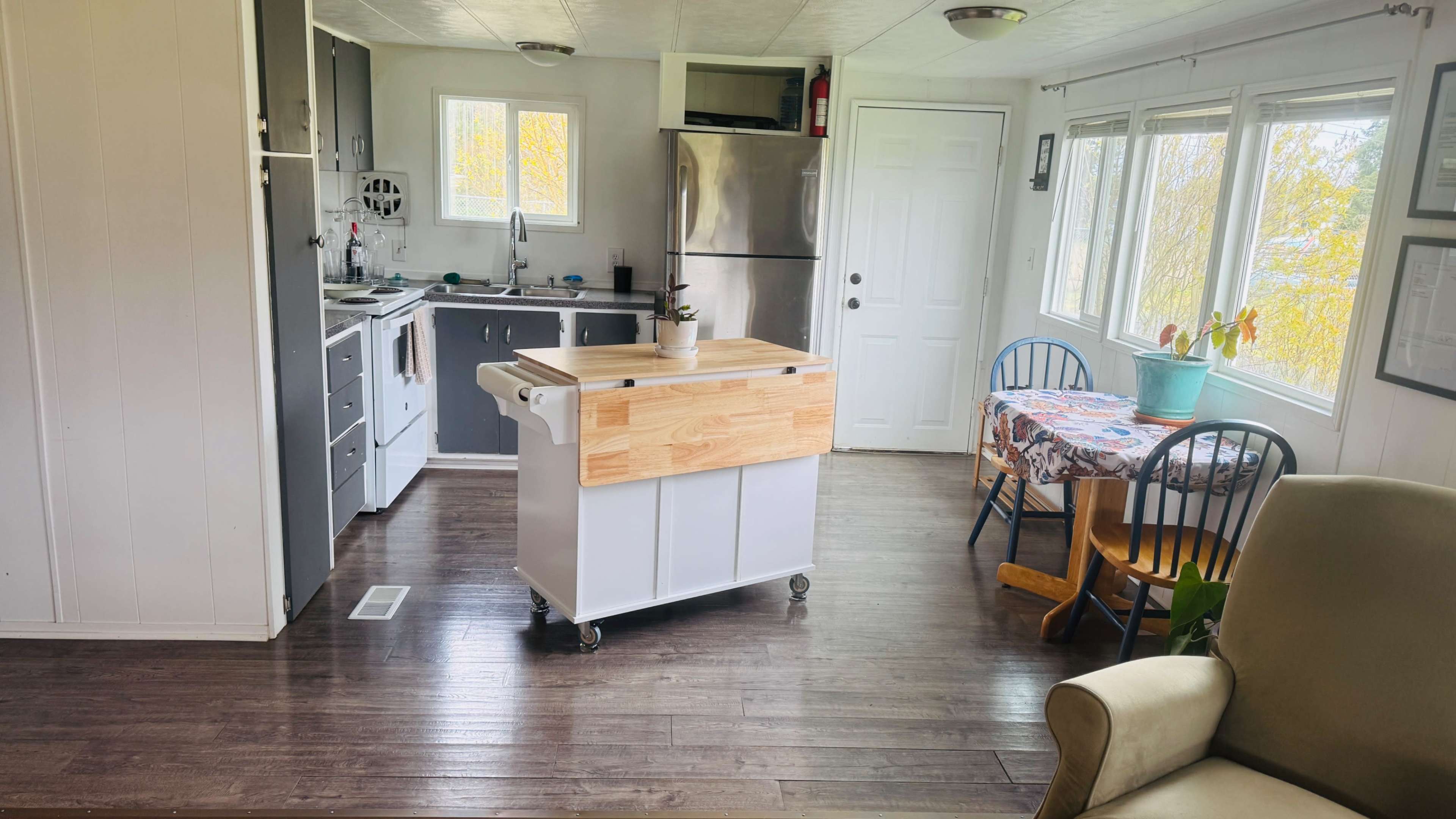 The image shows a compact kitchen and dining area featuring a central island with wheels, stainless steel appliances, and wooden furniture.