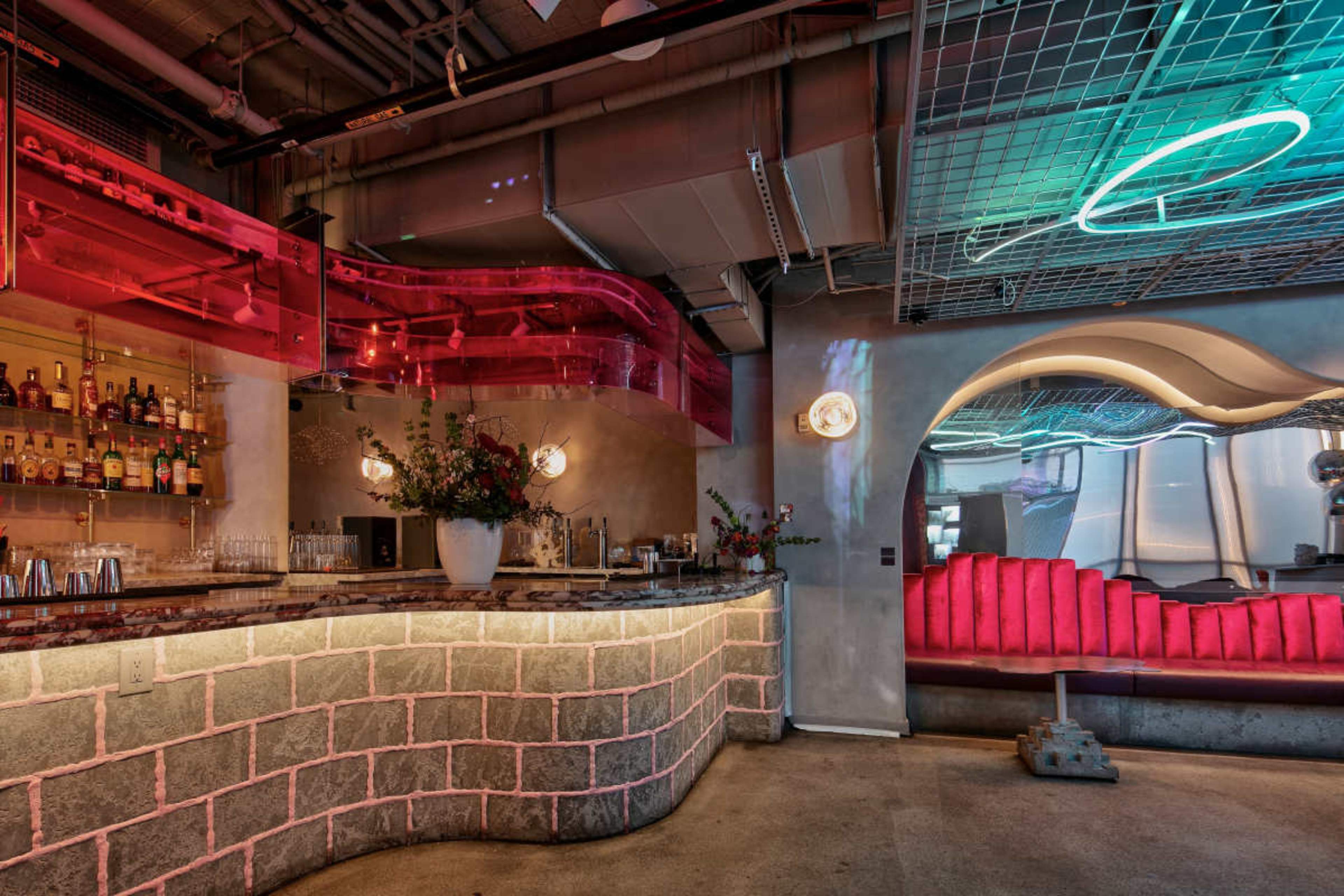 The image shows a modern bar interior featuring a curved, illuminated counter with a colorful lighting design and an abstract seating area in the background.