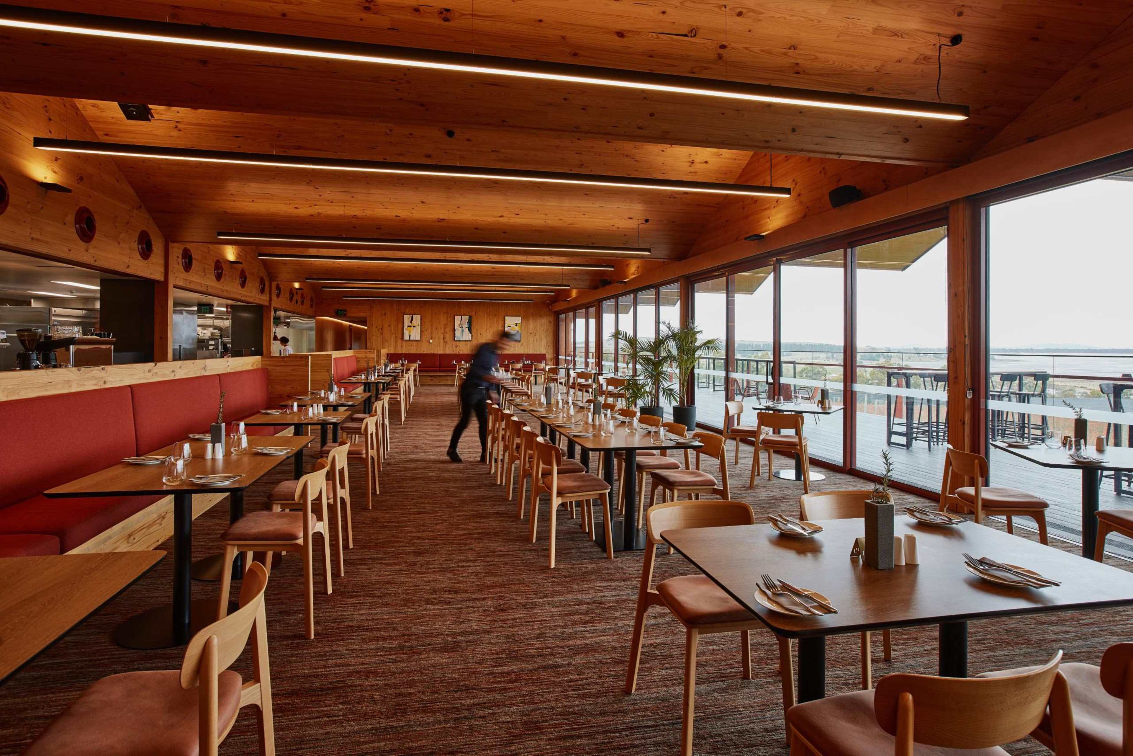 The interior of a modern restaurant features wooden beams and furnishings, with numerous tables and chairs arranged neatly, alongside large windows offering a view of the outdoors.