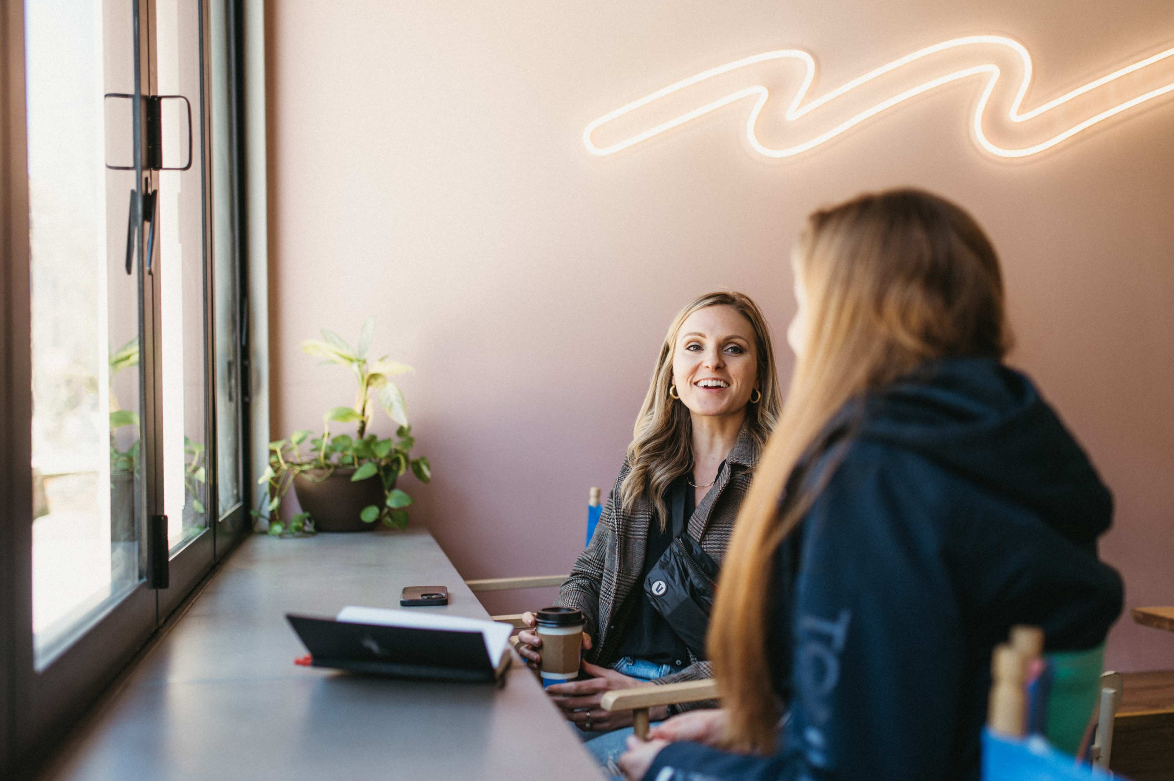 Two women are seated at a table in a café, engaged in conversation, with a laptop and a coffee cup on the table and a neon light on the wall behind them.