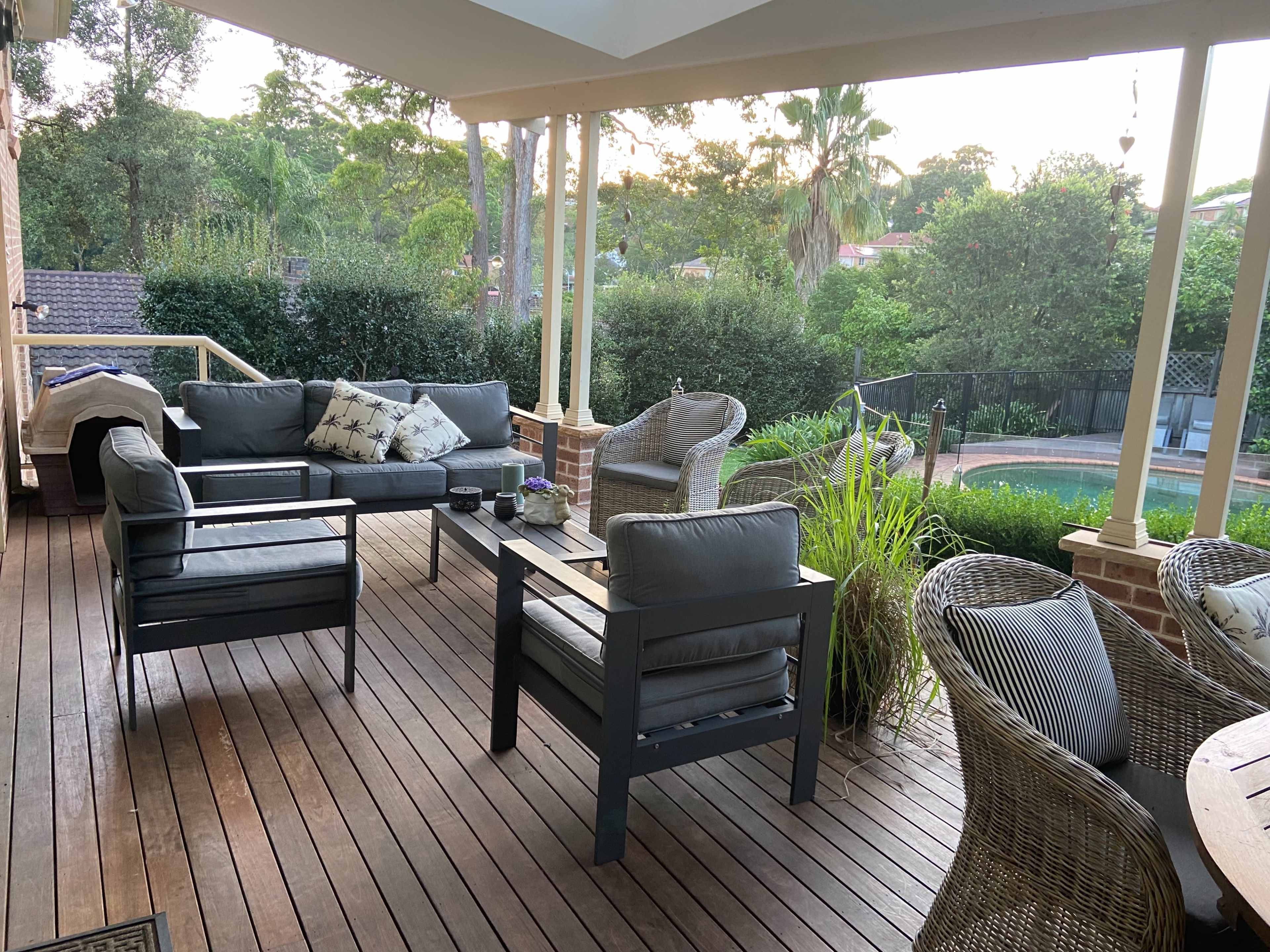 A patio features a seating area with gray couches and a wicker chair, surrounded by greenery and overlooking a swimming pool.