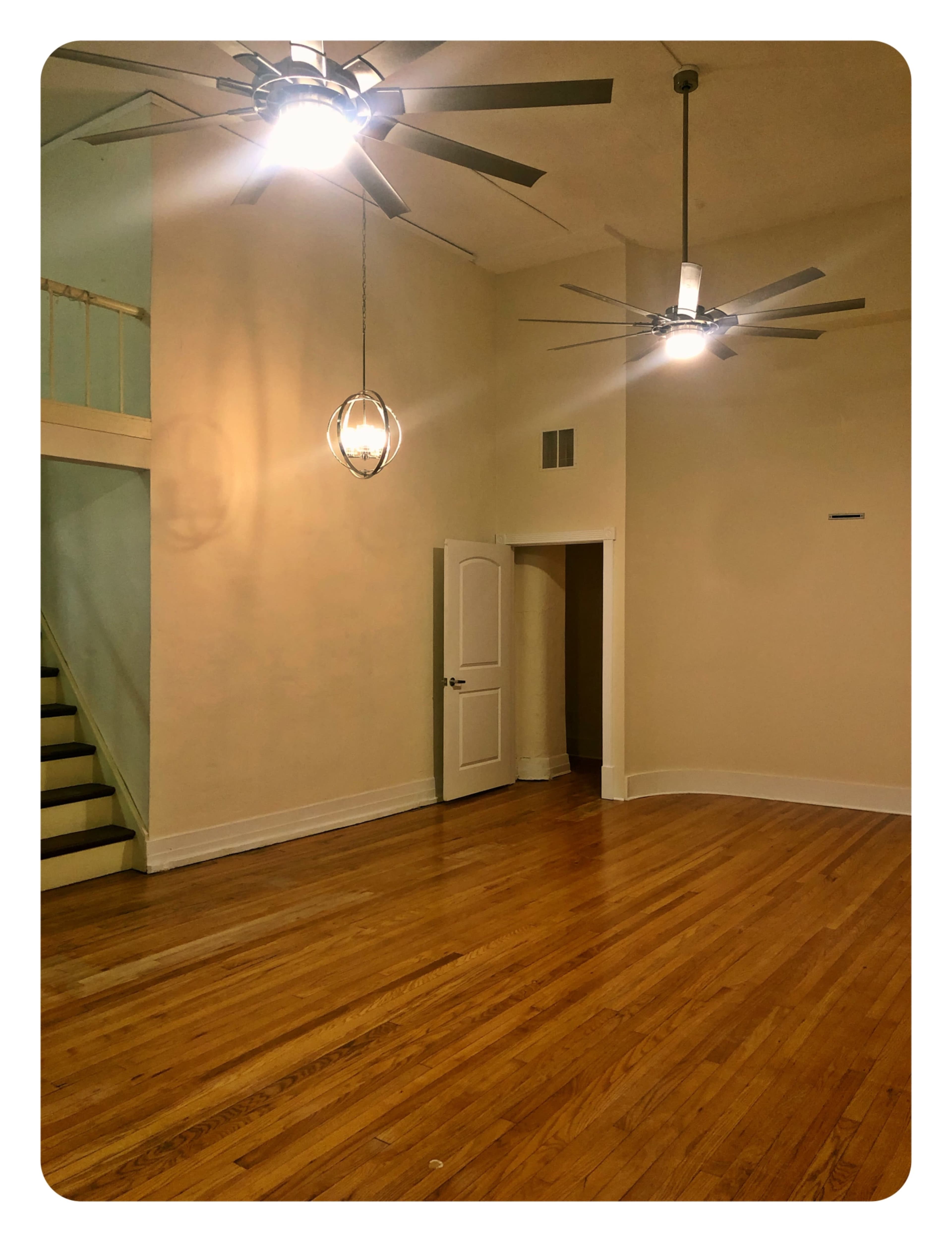 A spacious room features hardwood floors, two ceiling fans, and a doorway leading to a staircase.