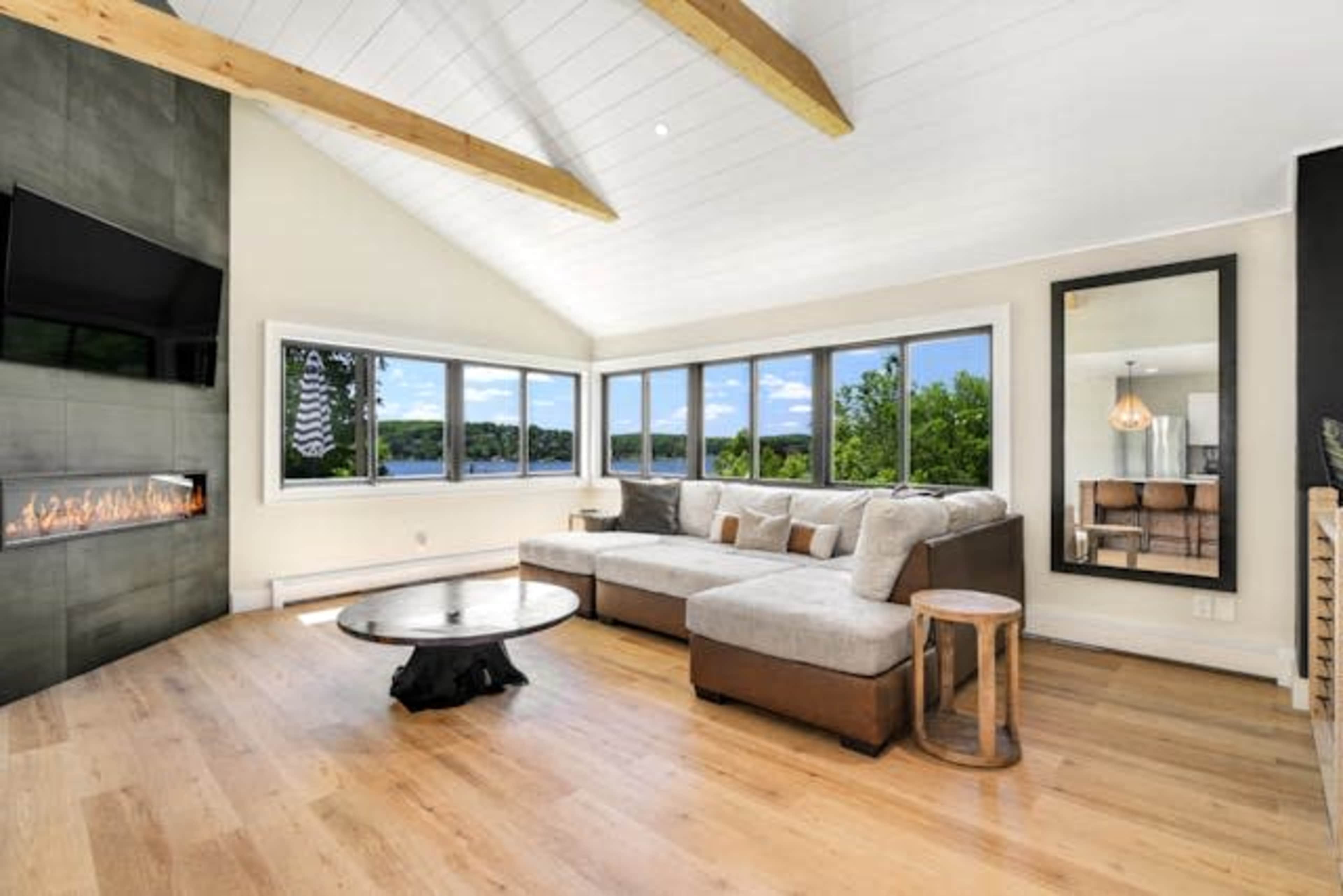 The image shows a modern living room with a large sectional sofa, a wall-mounted television, and a view of a lake through large windows.