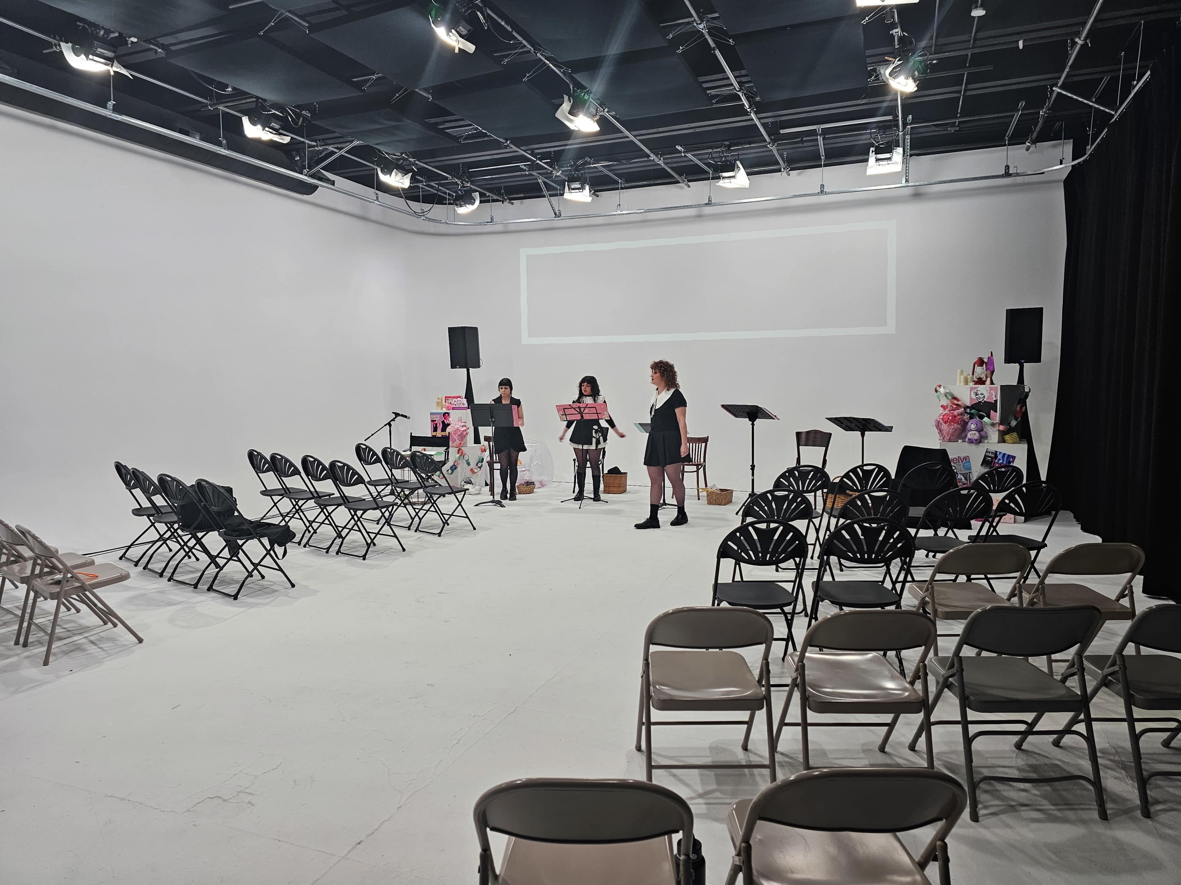 A spacious, empty studio is set up for an event, with chairs arranged in rows and a stage area where three women are preparing.
