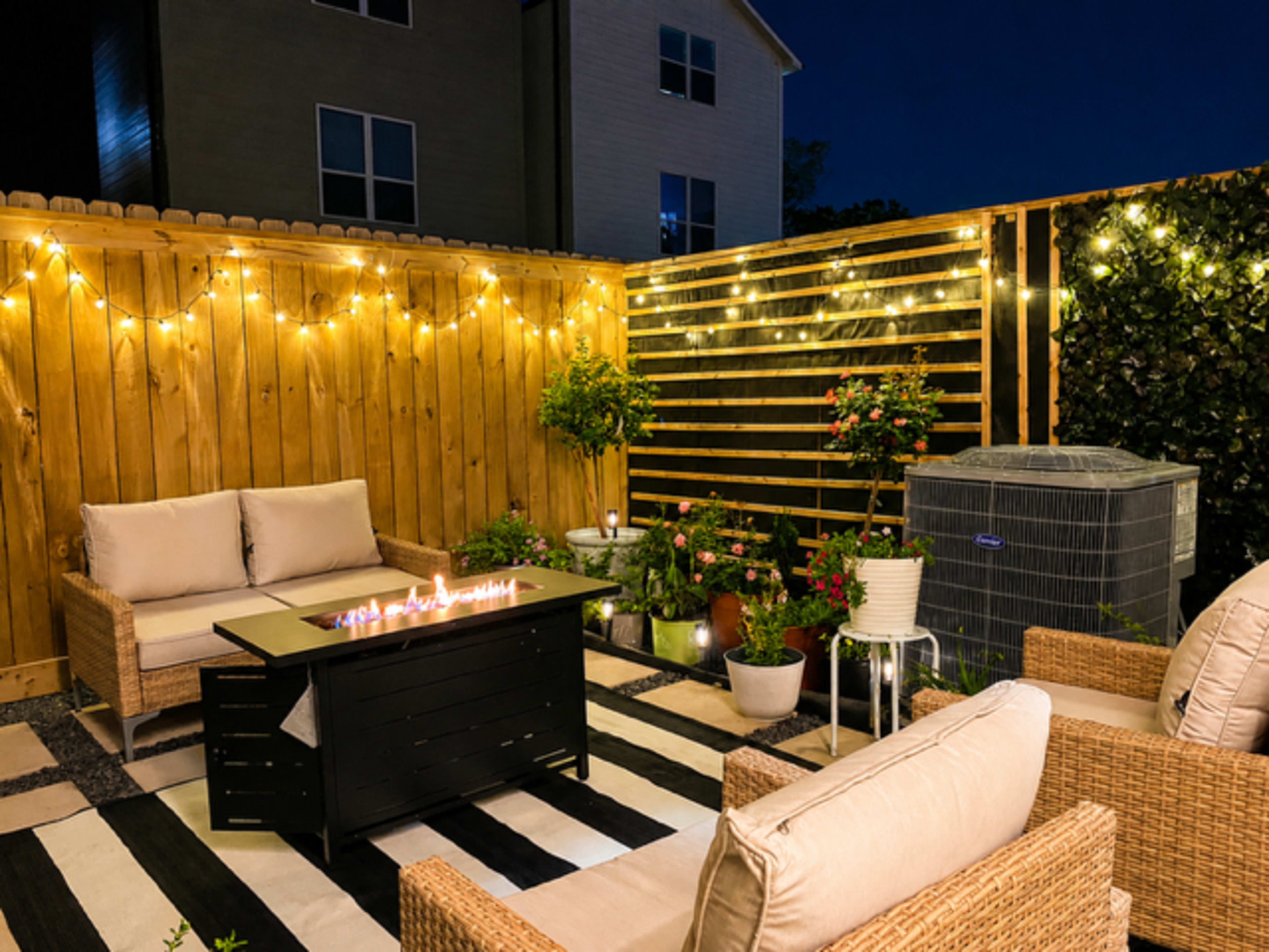 A cozy outdoor seating area is illuminated by string lights, featuring a fire table surrounded by woven chairs on a striped rug.