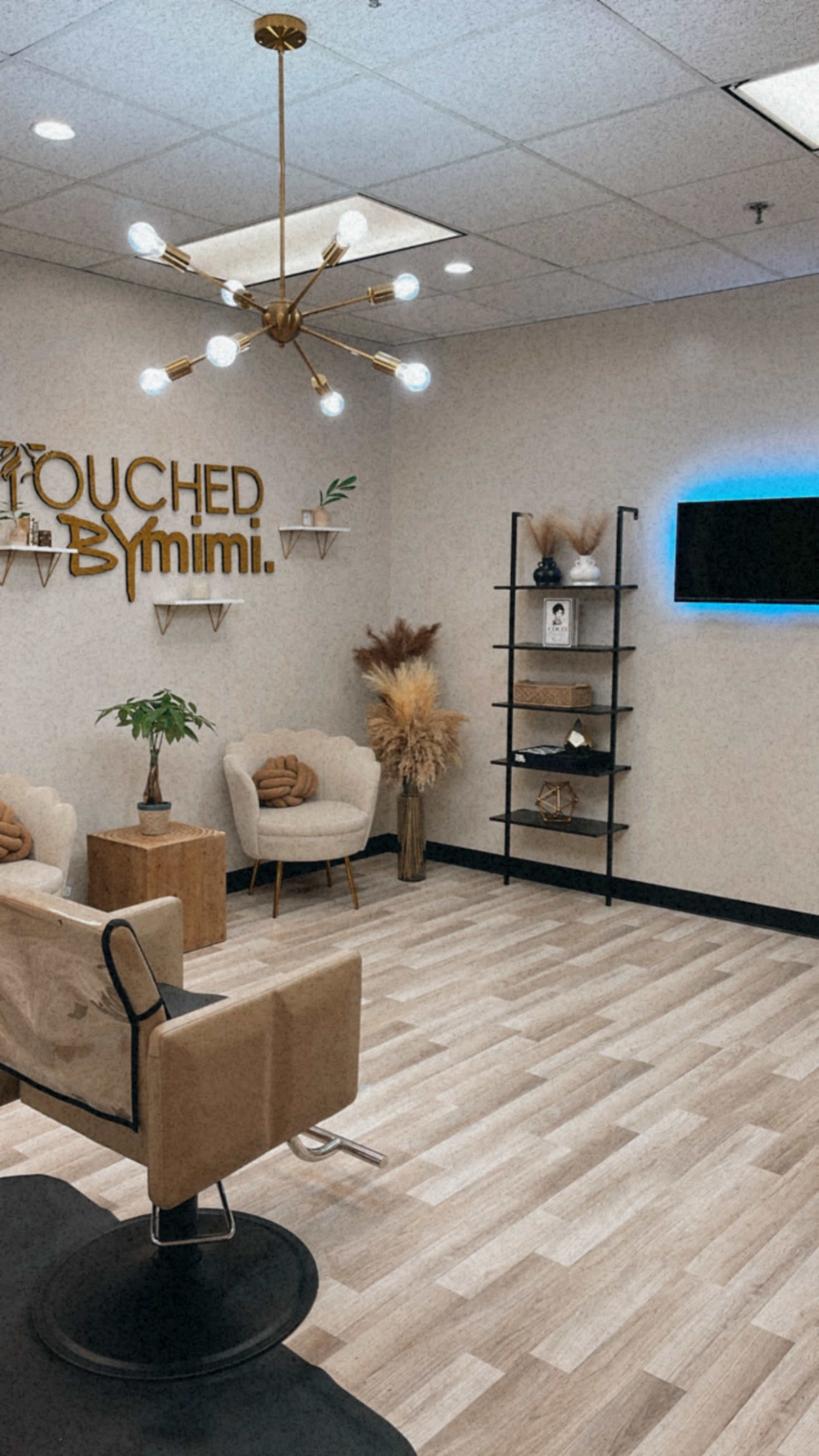 The image shows a modern salon interior with stylish seating, decorative plants, and a wall sign that reads "TOUCHED BY mimi."