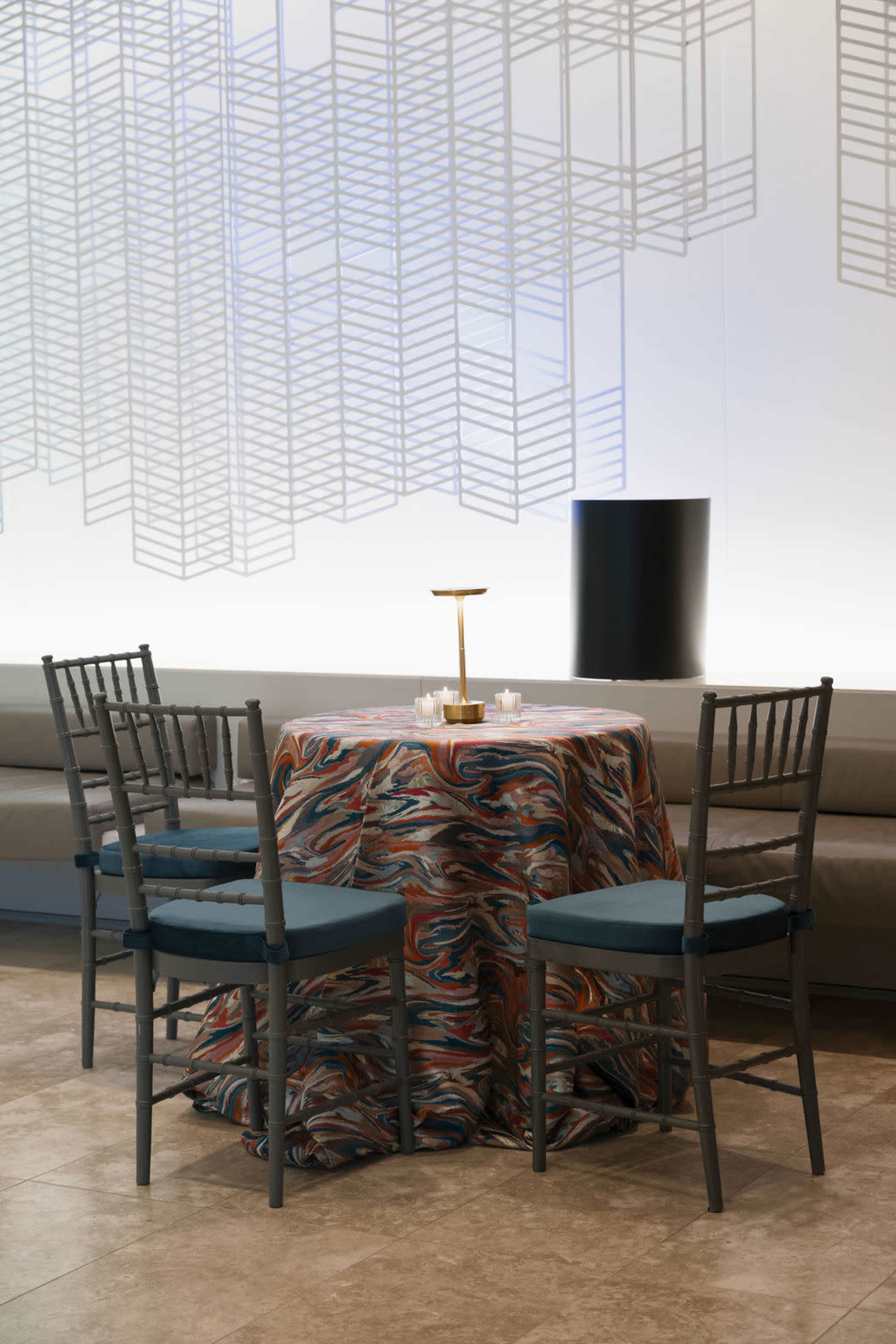A circular table covered with a patterned cloth is set with candles and surrounded by four chairs in a minimalist room with a modern design.