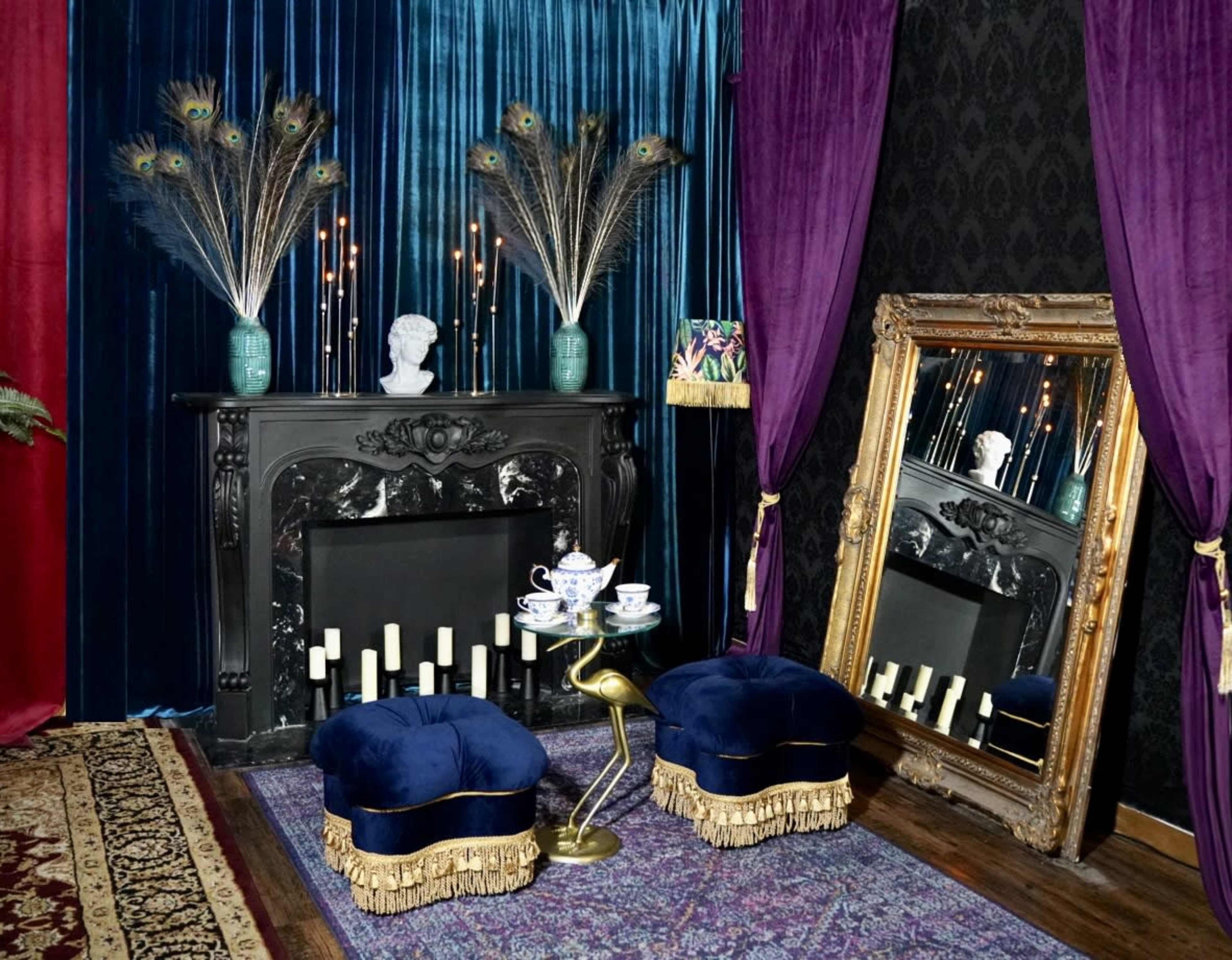 An ornate fireplace adorned with candles and a bust, flanked by peacock feathers in vases, with a large mirror and two velvet stools in front.