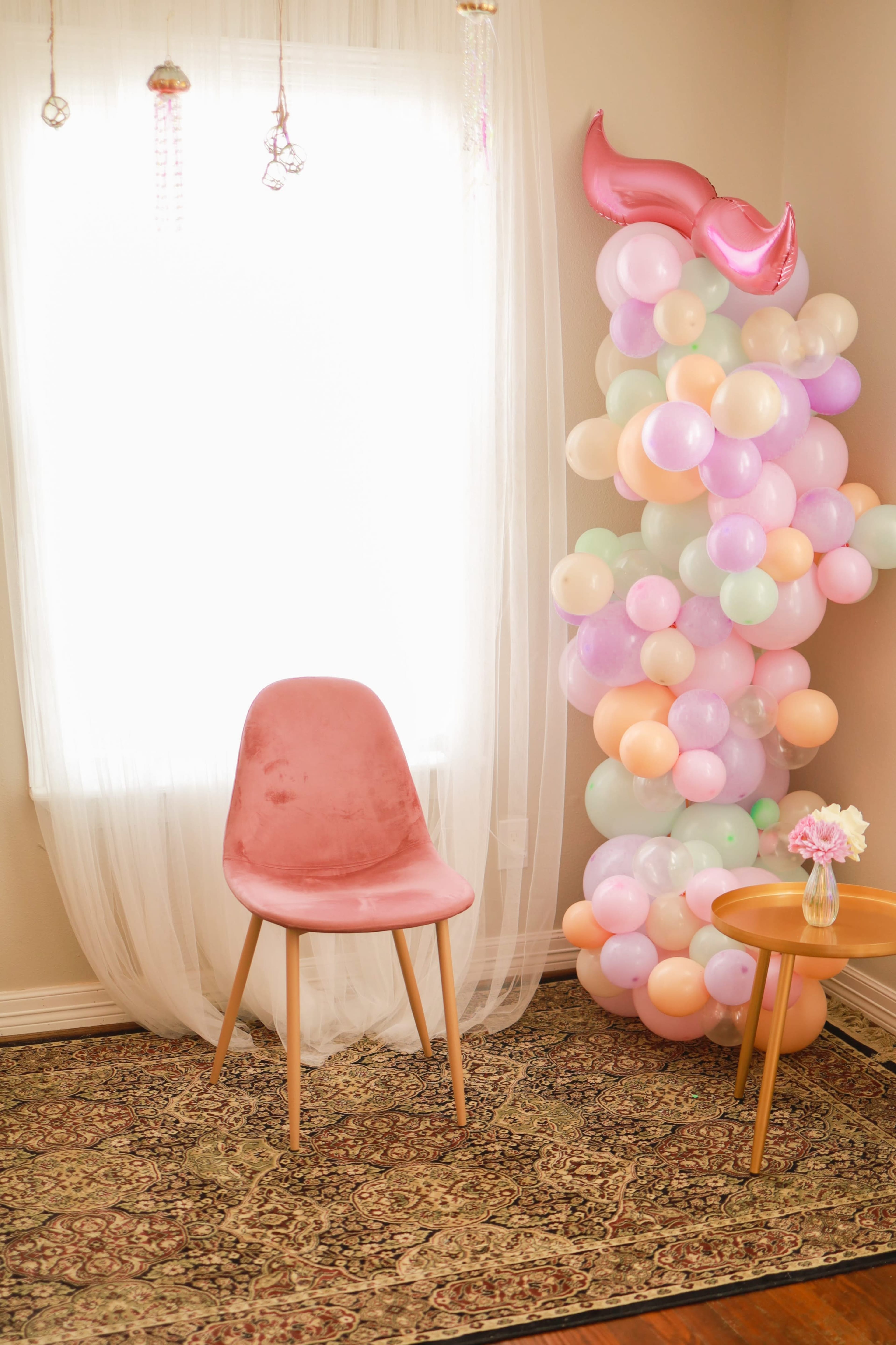 A pink chair stands next to a colorful balloon arrangement in a softly lit room with a window draped in sheer curtains.