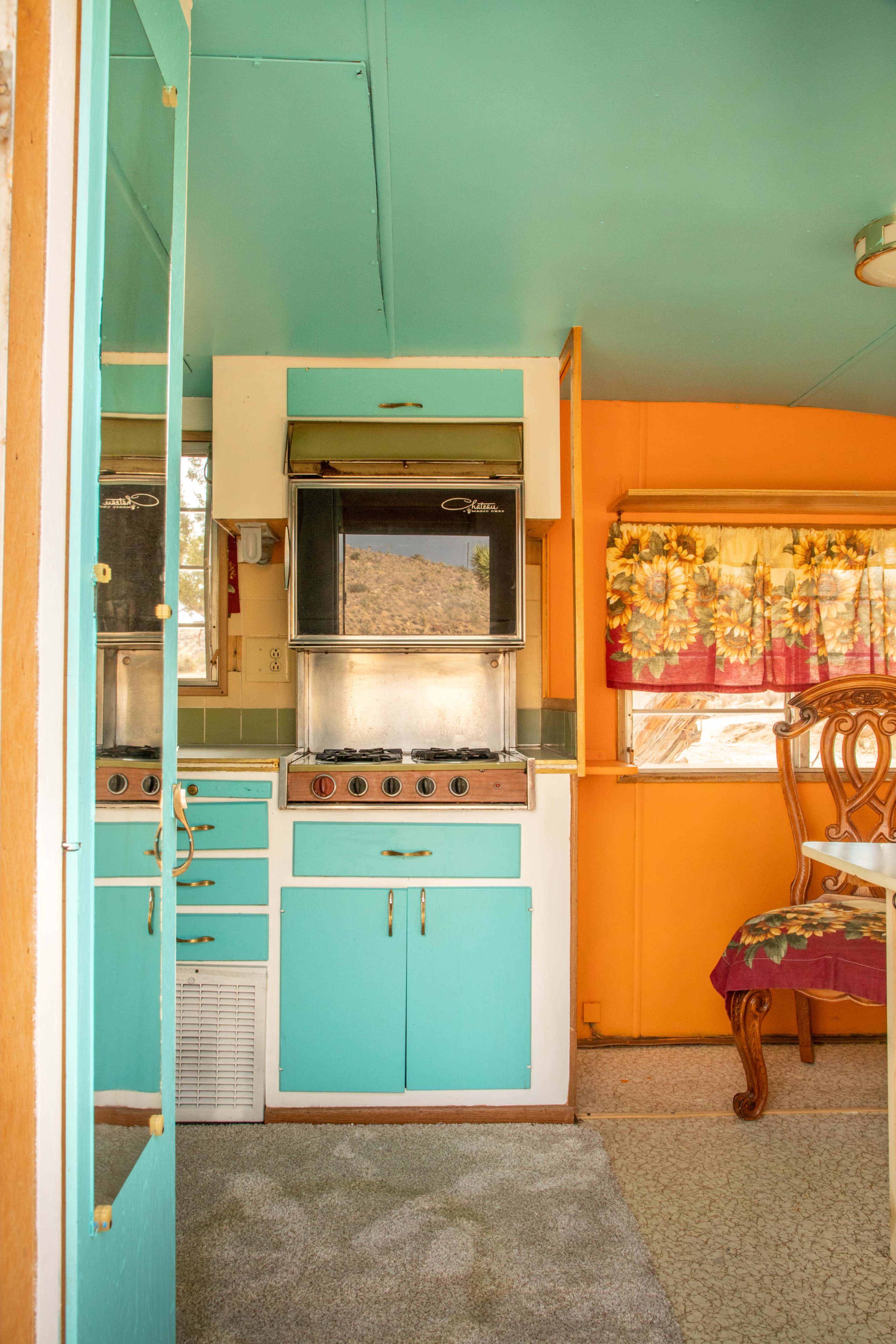 A small kitchen with turquoise and orange cabinetry, a vintage stove, and a dining area with a chair near a window.