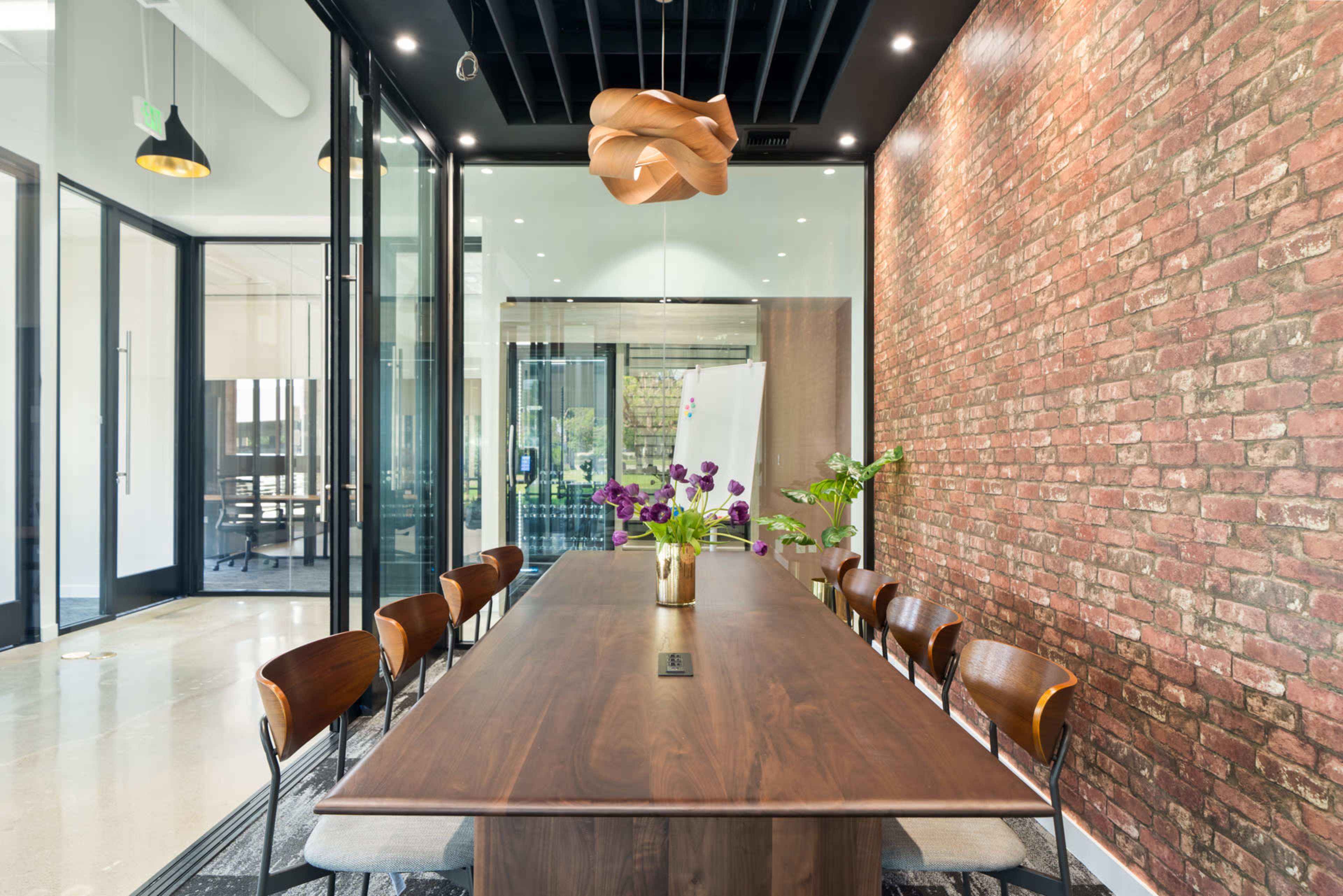 A long wooden conference table, surrounded by eight chairs, is set in a room with a brick wall and glass panels, featuring a decorative light fixture above and a vase of flowers at the center.