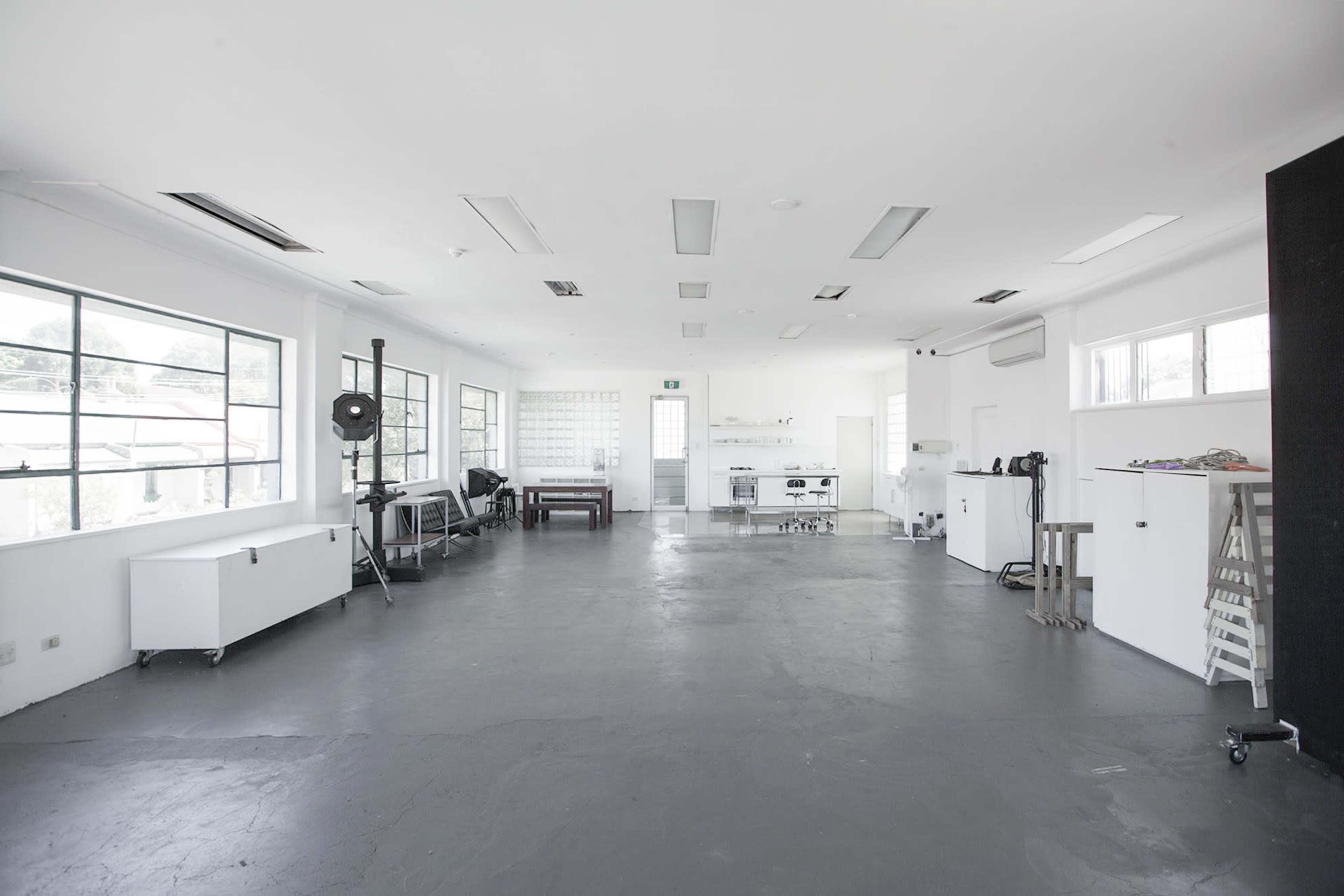 The image shows a spacious, minimalist studio with white walls, large windows, and various furniture items scattered throughout the floor.