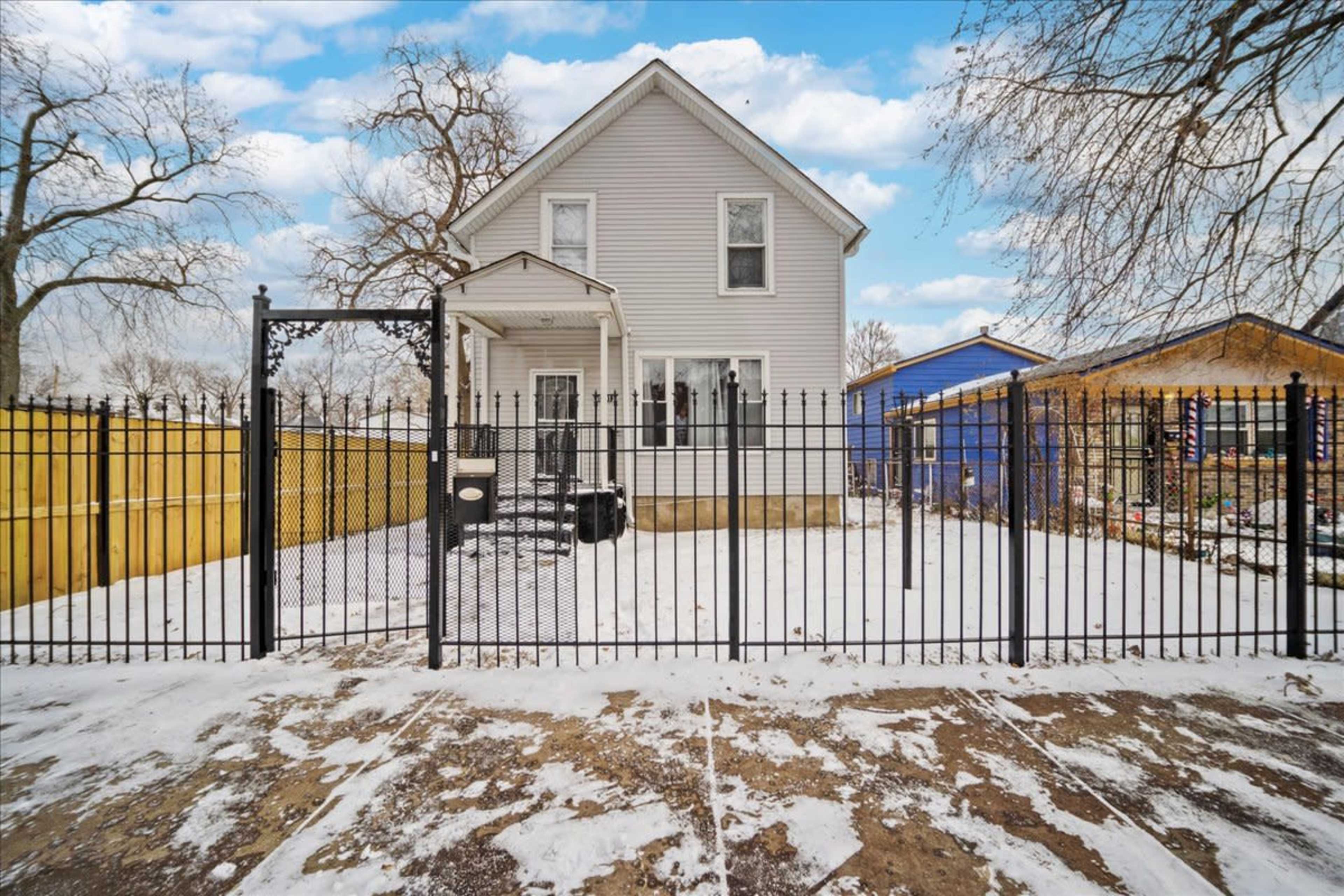 A two-story house with a fenced yard, covered patio, and snow-covered ground is surrounded by trees and neighboring structures.