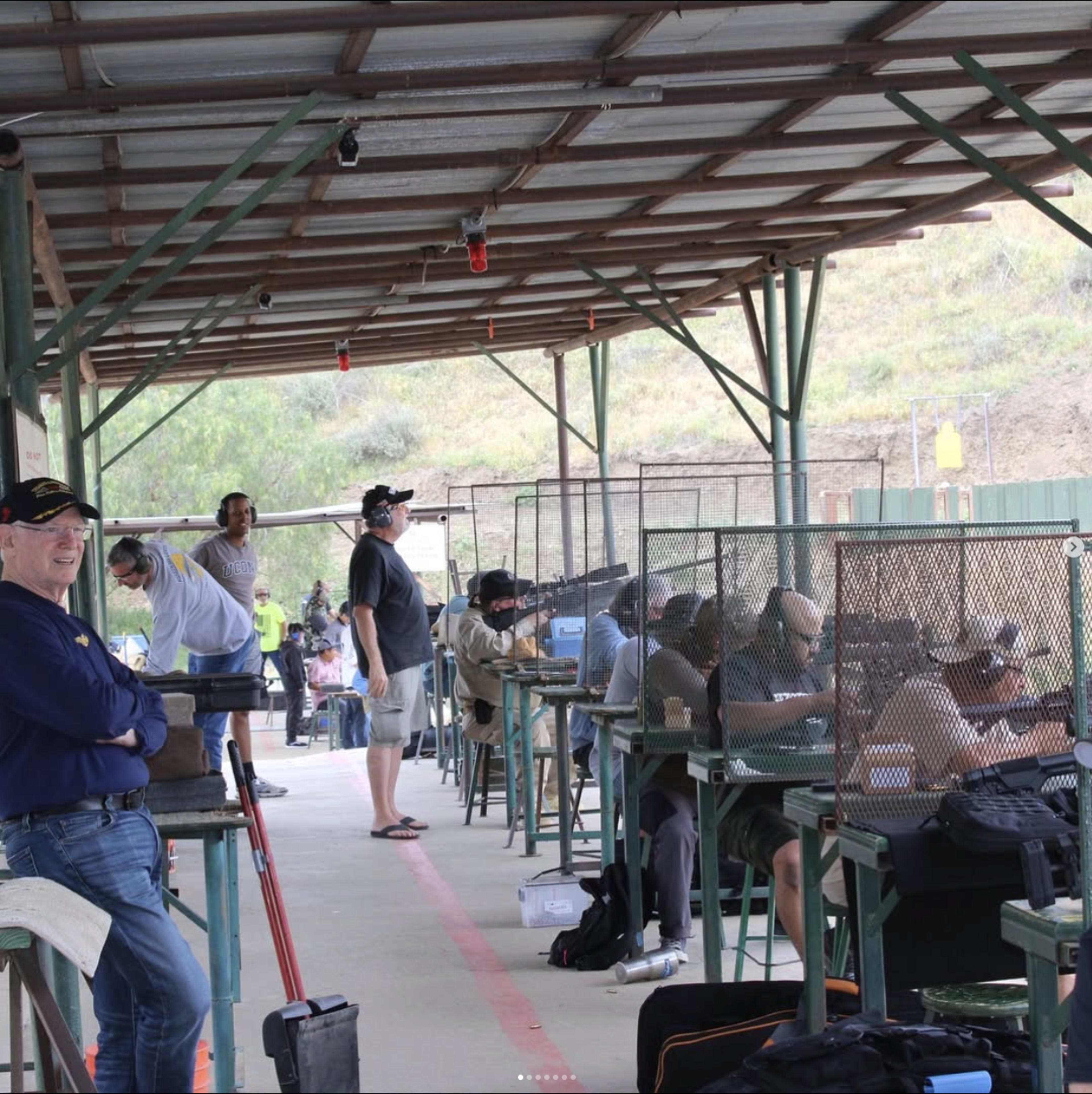 A group of people is participating in a shooting range activity, with several individuals stationed at benches behind protective mesh.