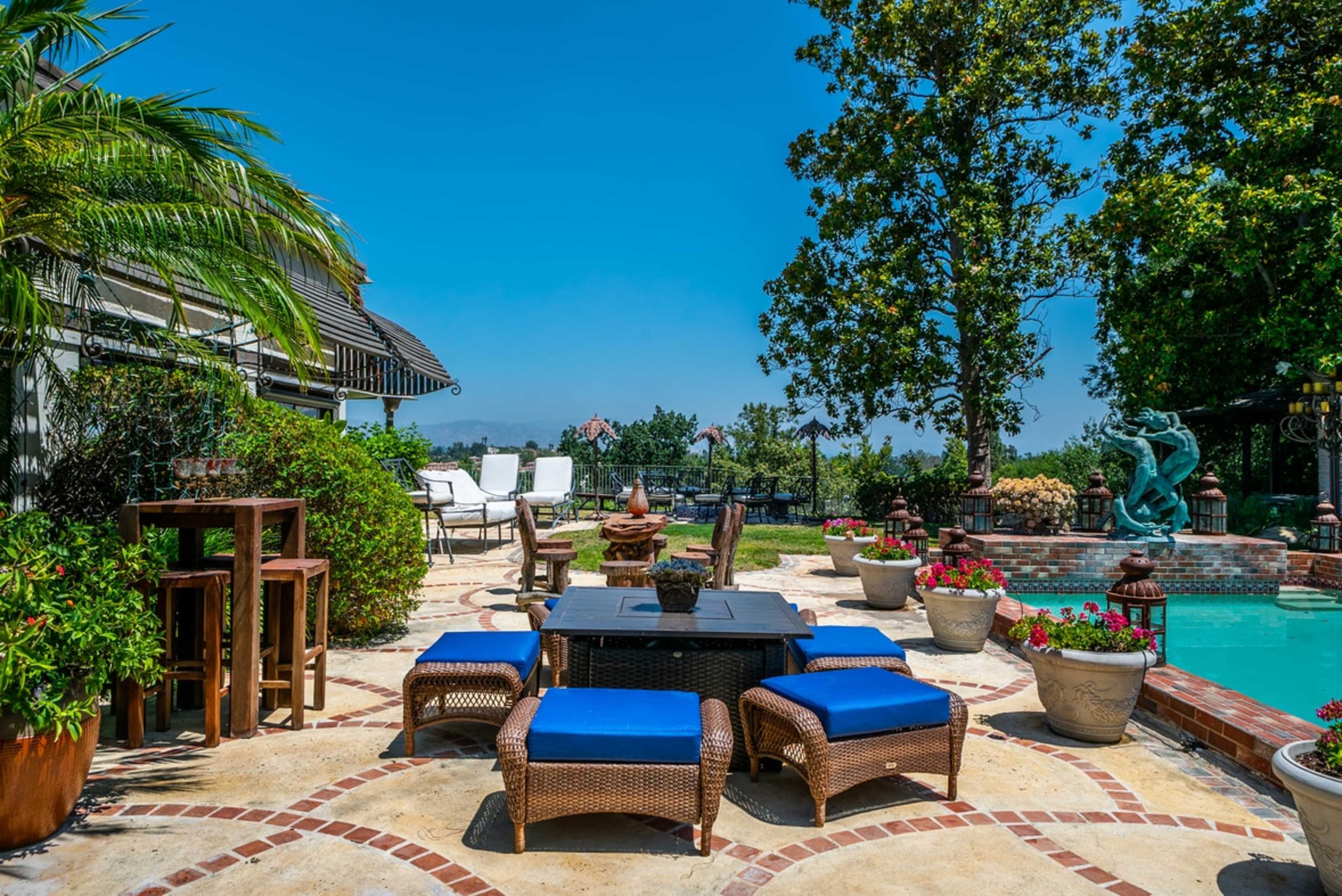 The image shows a well-furnished patio with blue cushioned chairs and a table near a swimming pool, surrounded by potted plants and a scenic view.
