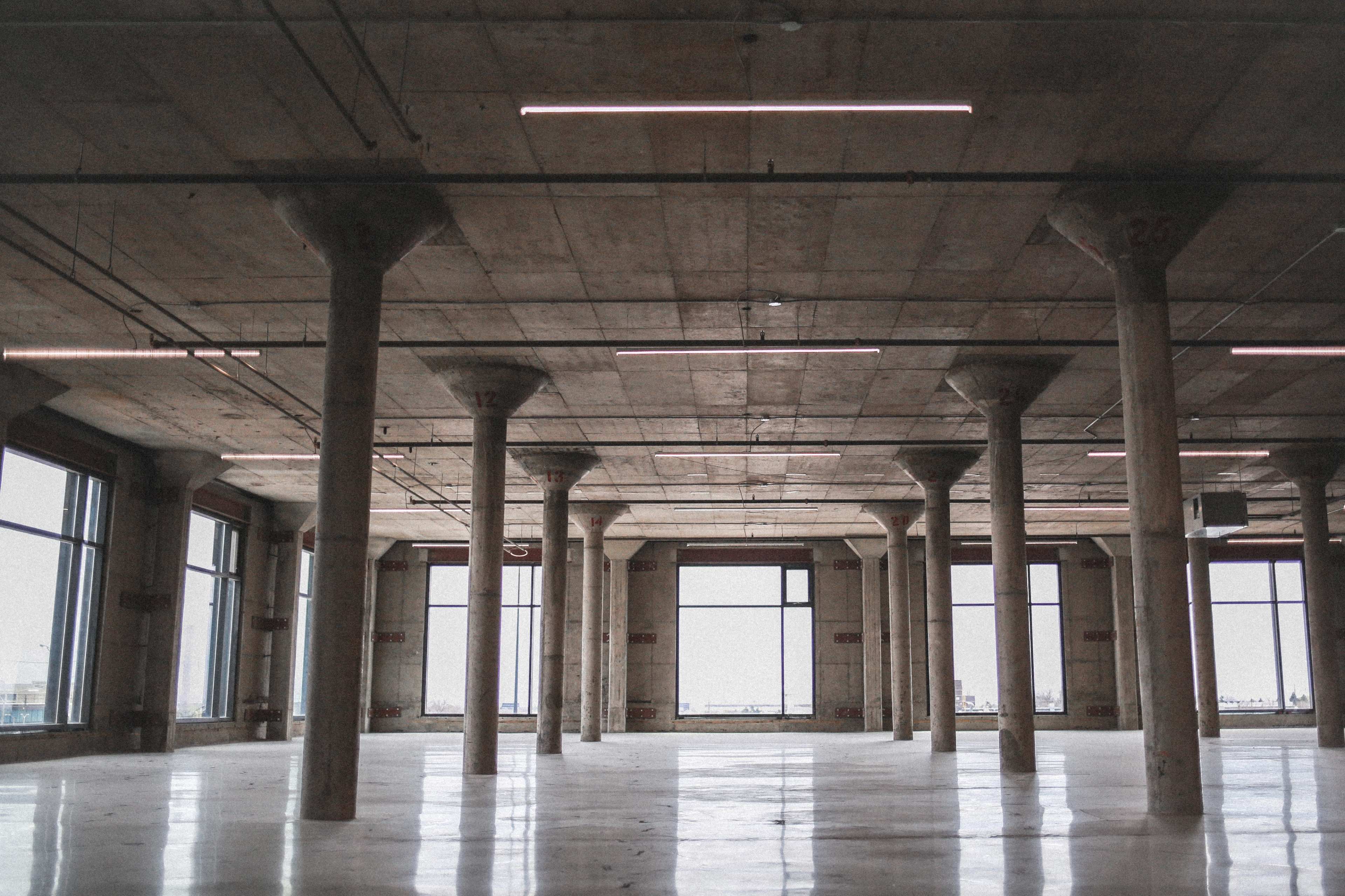 The image shows a spacious, unfinished interior of a building with large windows and exposed concrete pillars.