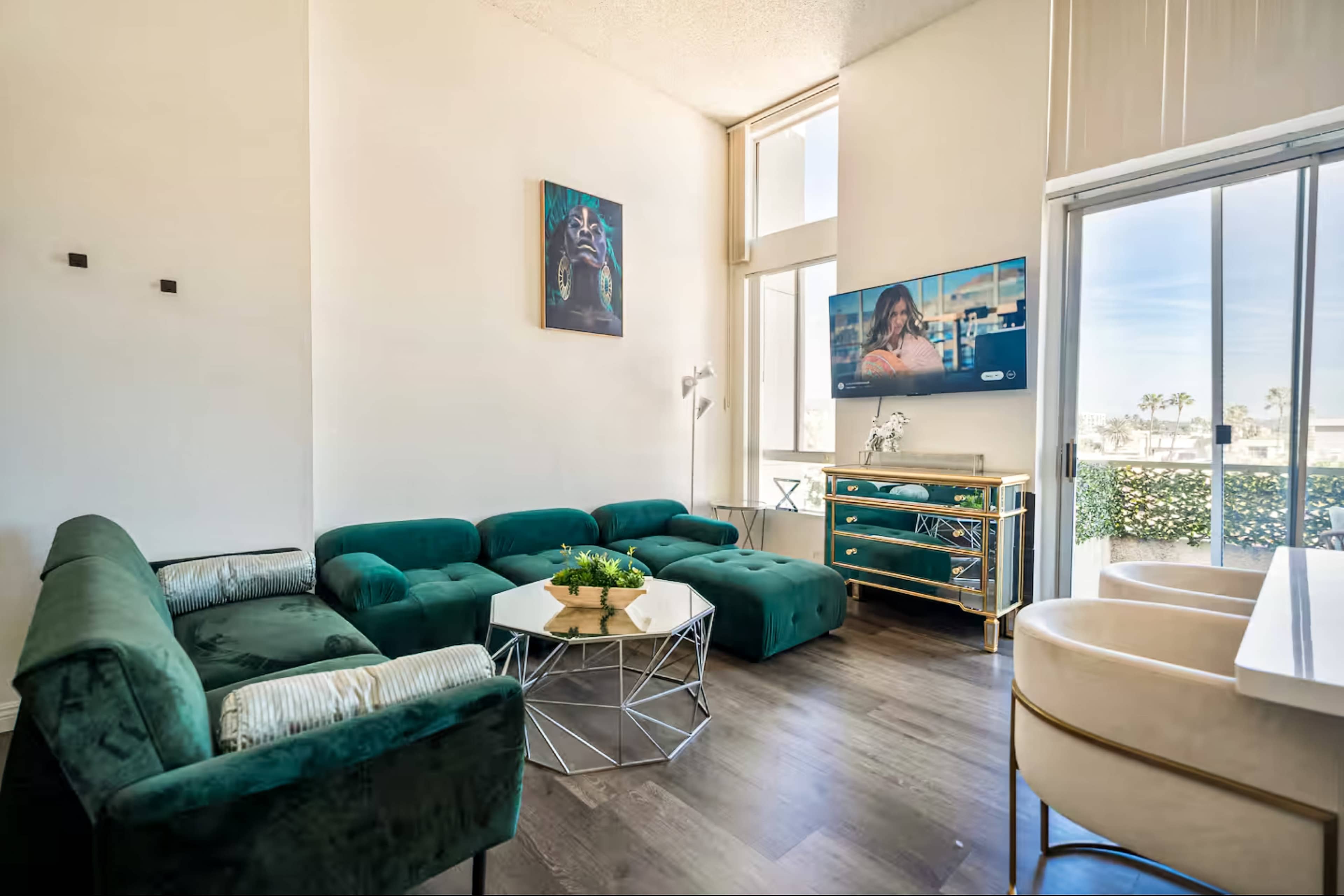 A modern living room features a green sectional sofa, a glass coffee table, a decorative console with a television above it, and large windows allowing natural light.