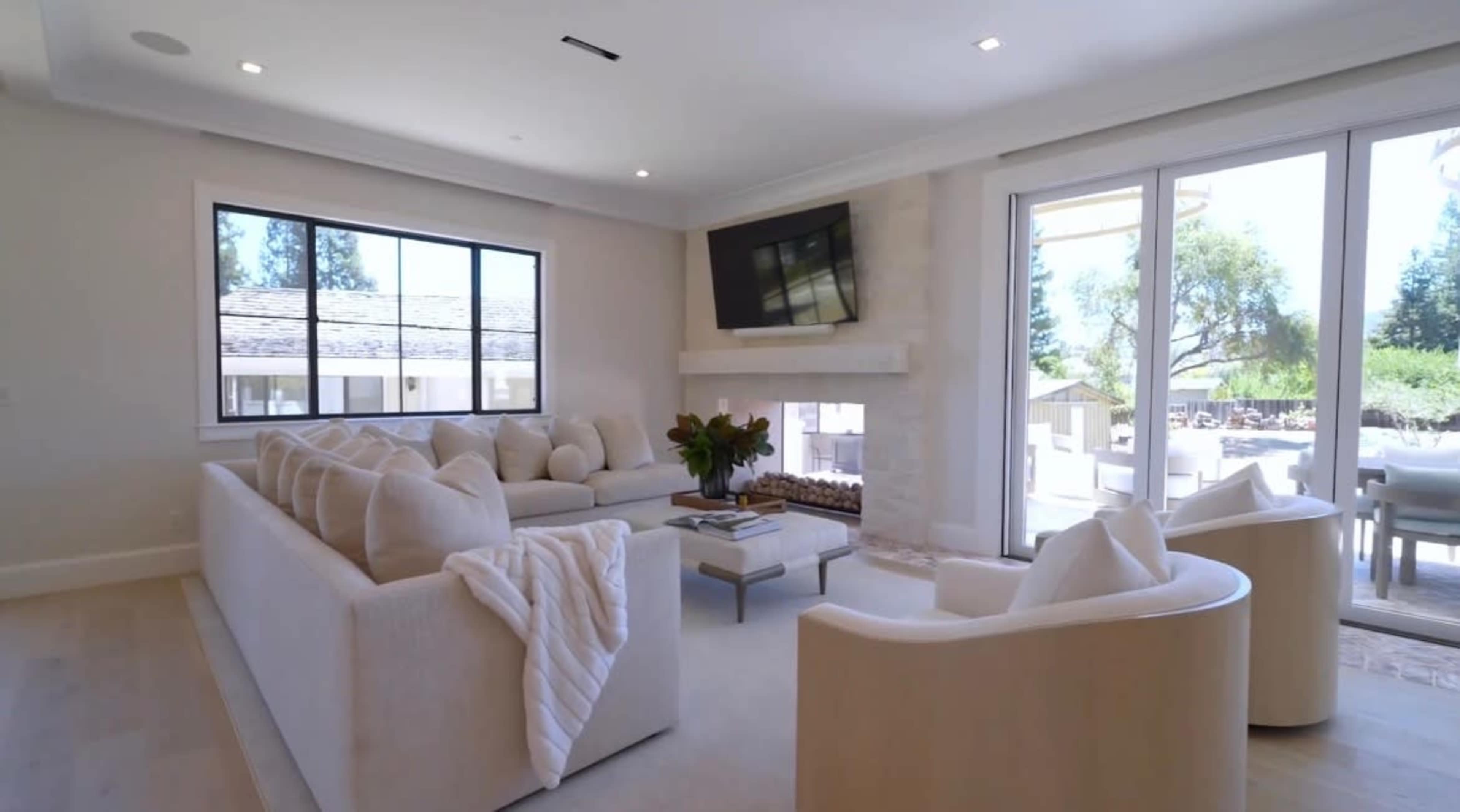 The image shows a modern living room with a large sofa, two armchairs, a television mounted on the wall, and large windows providing natural light.