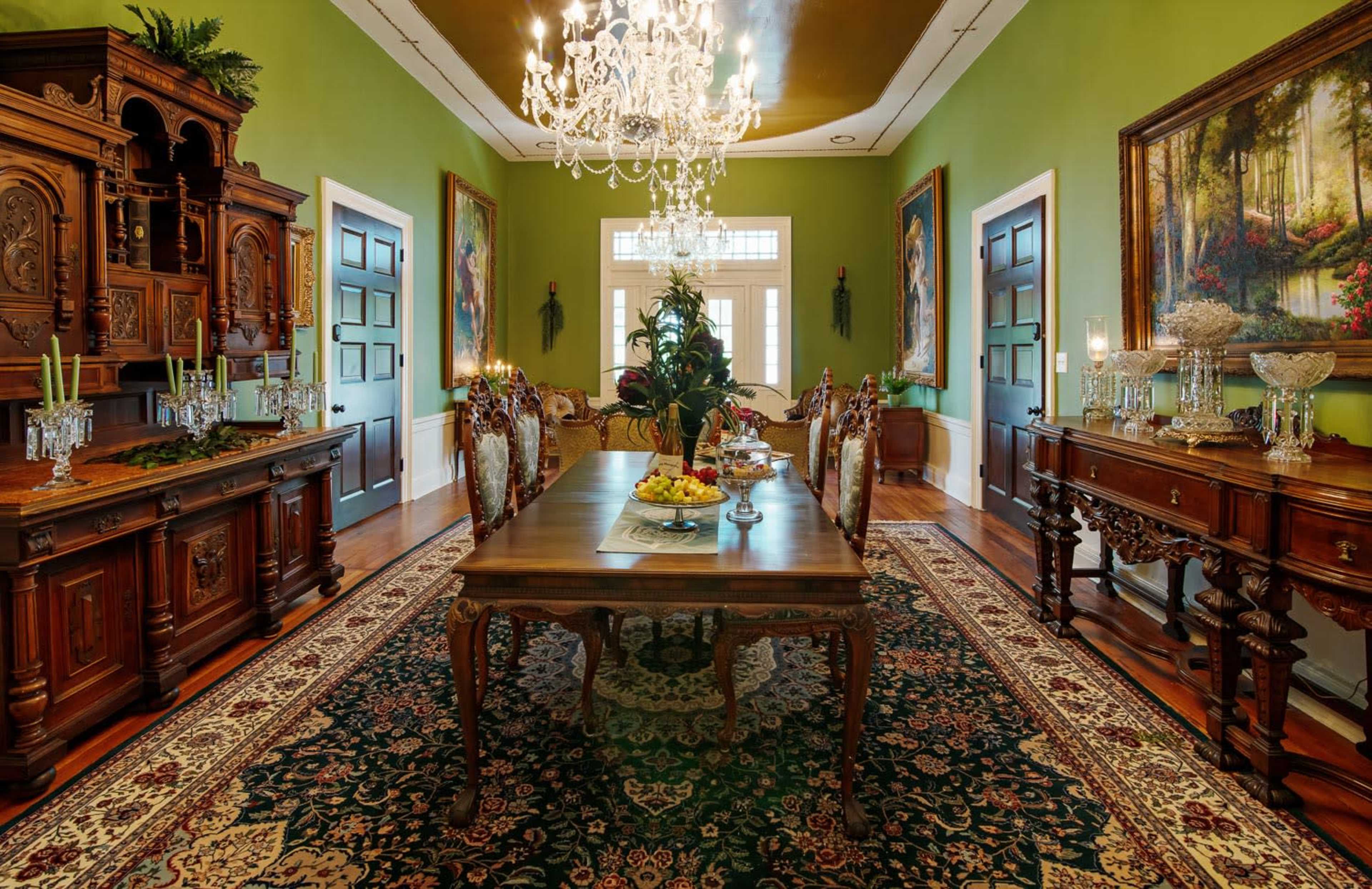 A dining room featuring a wooden table with a fruit centerpiece, surrounded by ornate chairs, and decorated with chandeliers and large paintings on green walls.