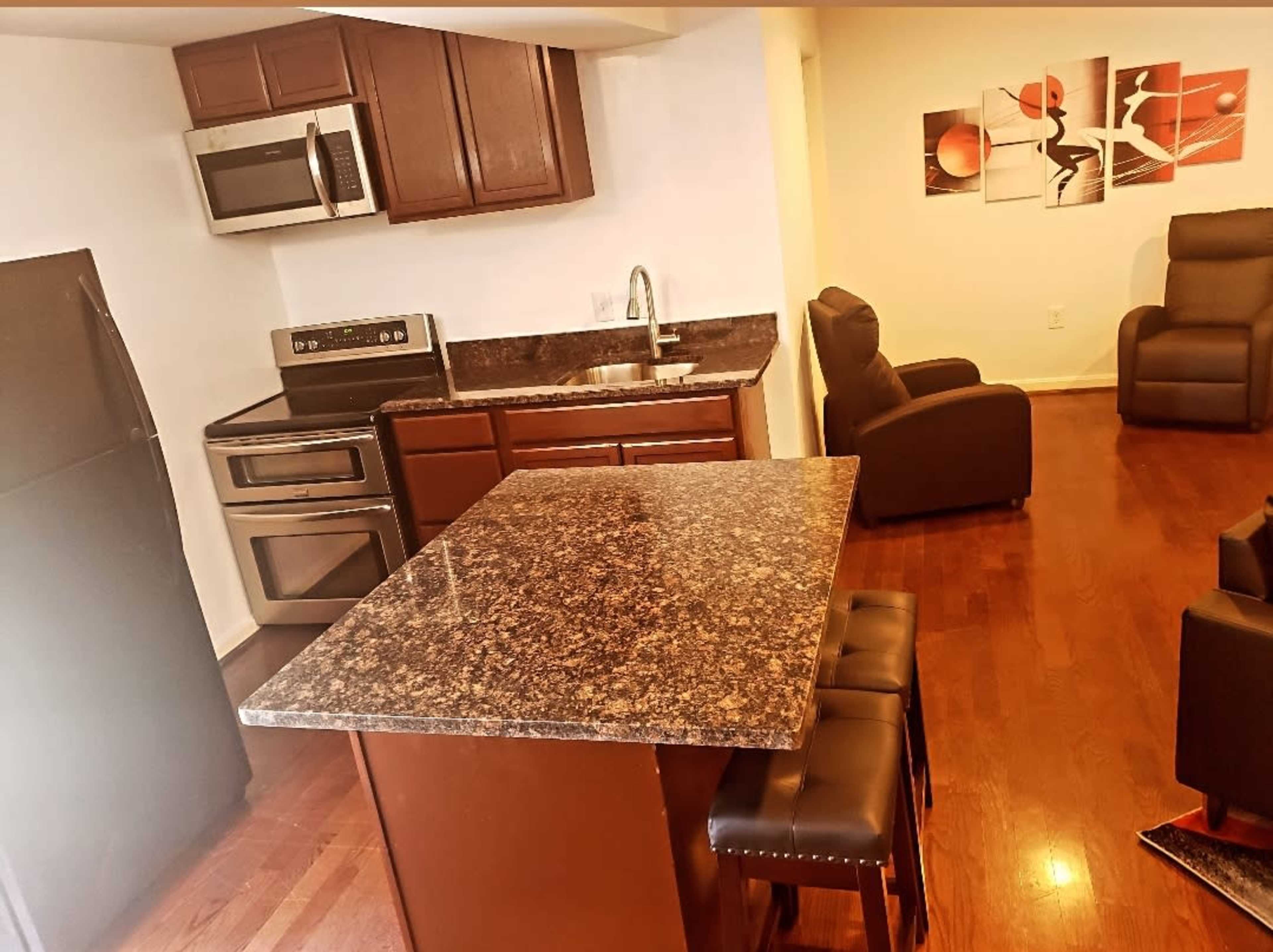 A kitchen with dark wood cabinets, stainless steel appliances, and a granite countertop island, connected to a living area with two black armchairs.