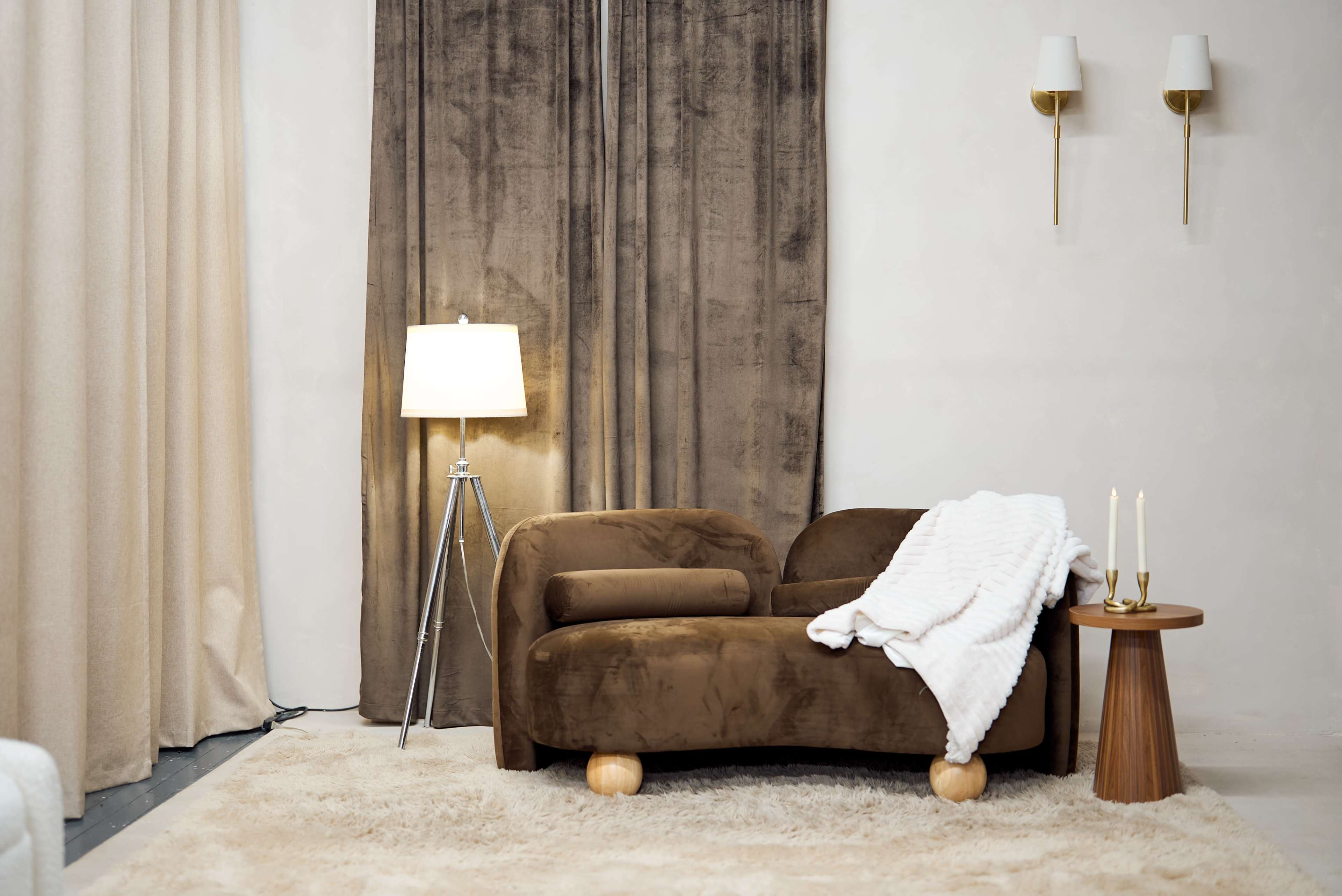 The scene features a cozy living space with a brown chaise lounge, a floor lamp, light-colored curtains, and a small wooden side table holding candles.