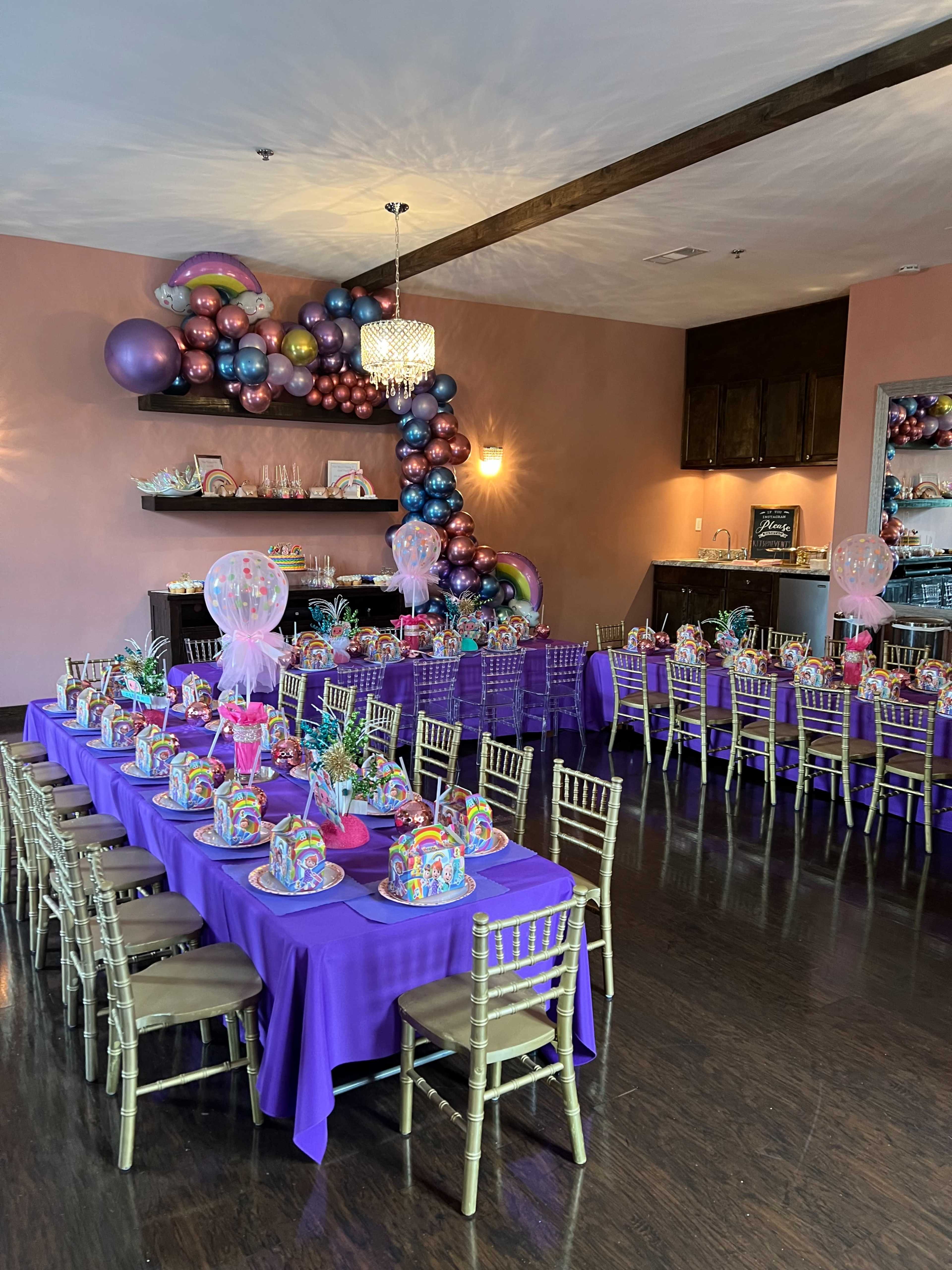 A banquet hall is set with two long tables covered in purple cloth, decorated with colorful centerpieces and balloons on one side of the room.
