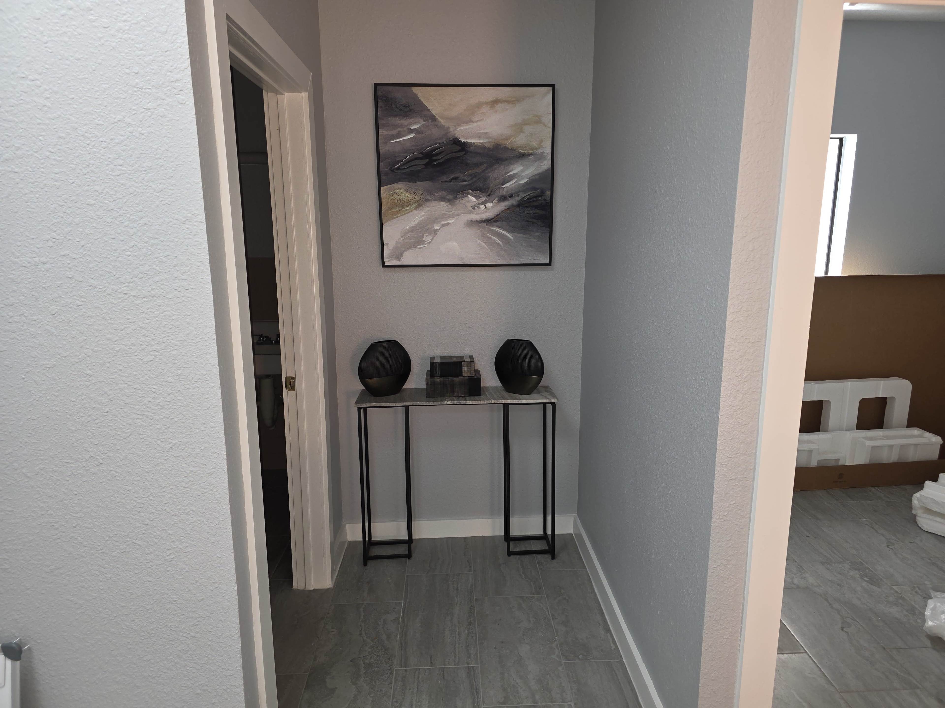The image shows a minimalist hallway leading to a bathroom, with a wall painting and a small table featuring decorative items.