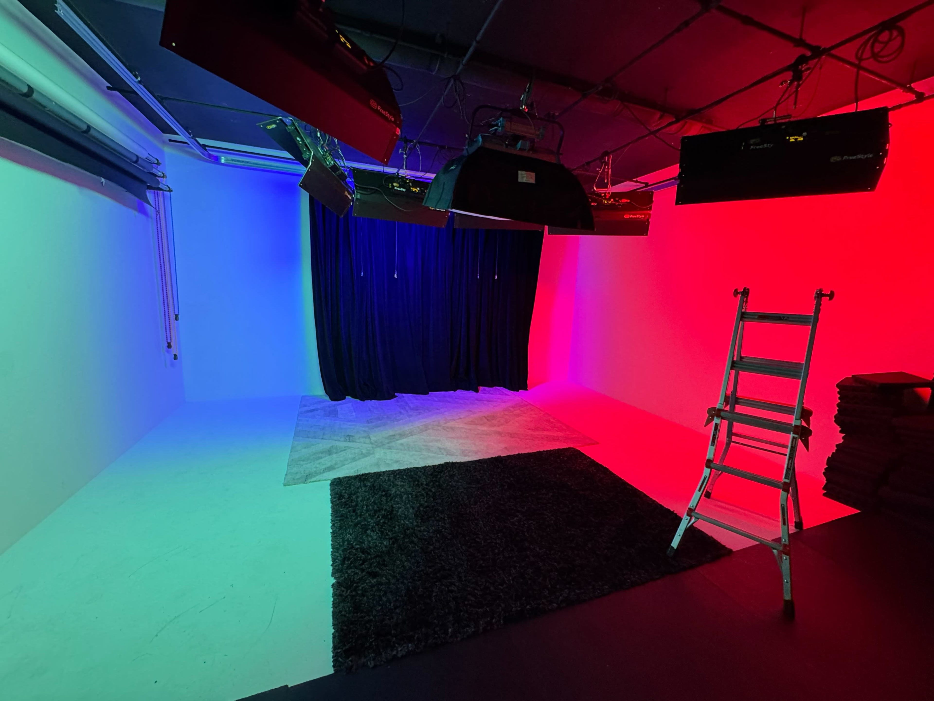 The image shows a studio space with colorful lighting, a black curtain, a ladder, and a rug on the floor.