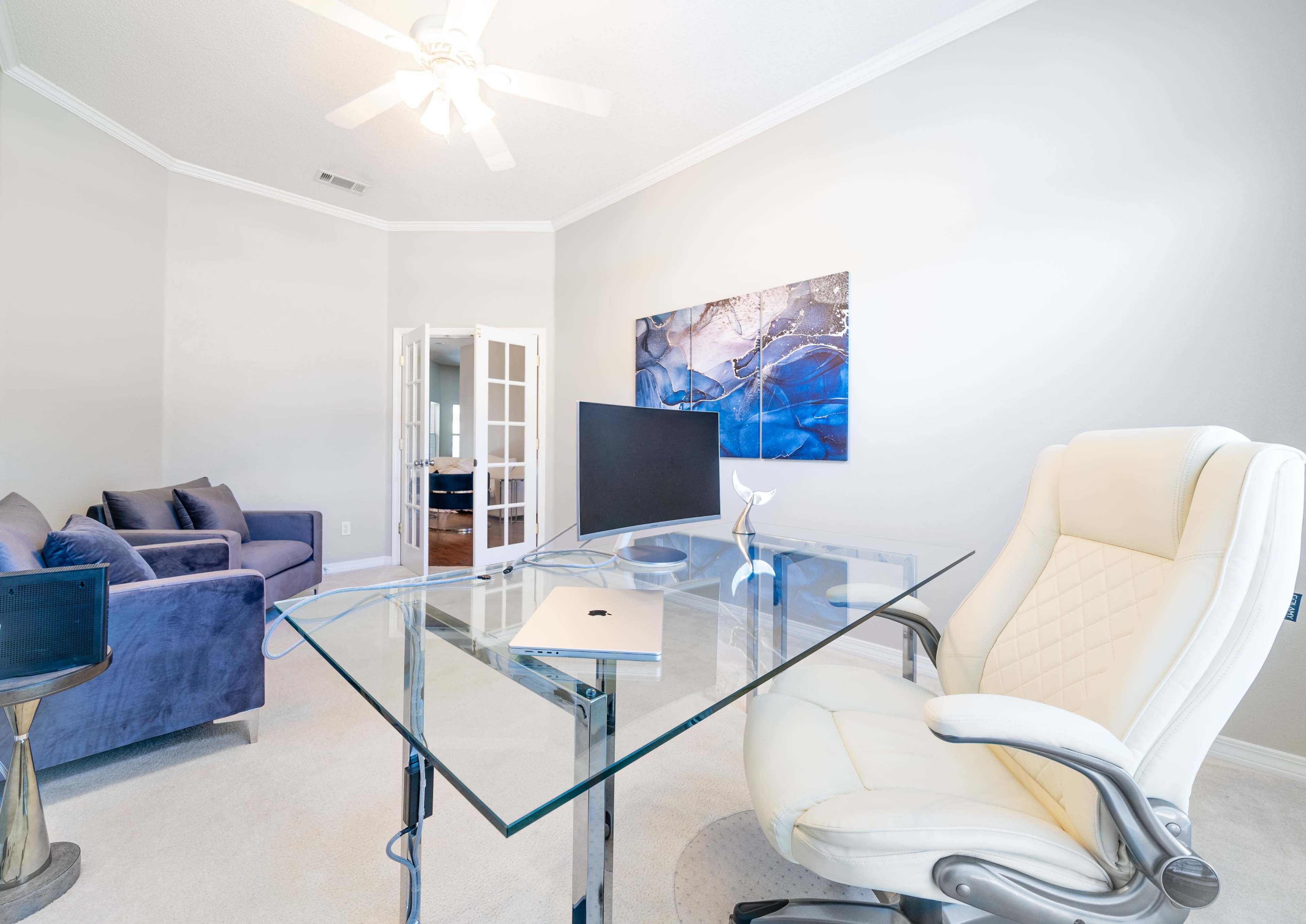 A modern office space features a clear glass desk and a white ergonomic chair, with a blue couch in the corner and a painting on the wall.