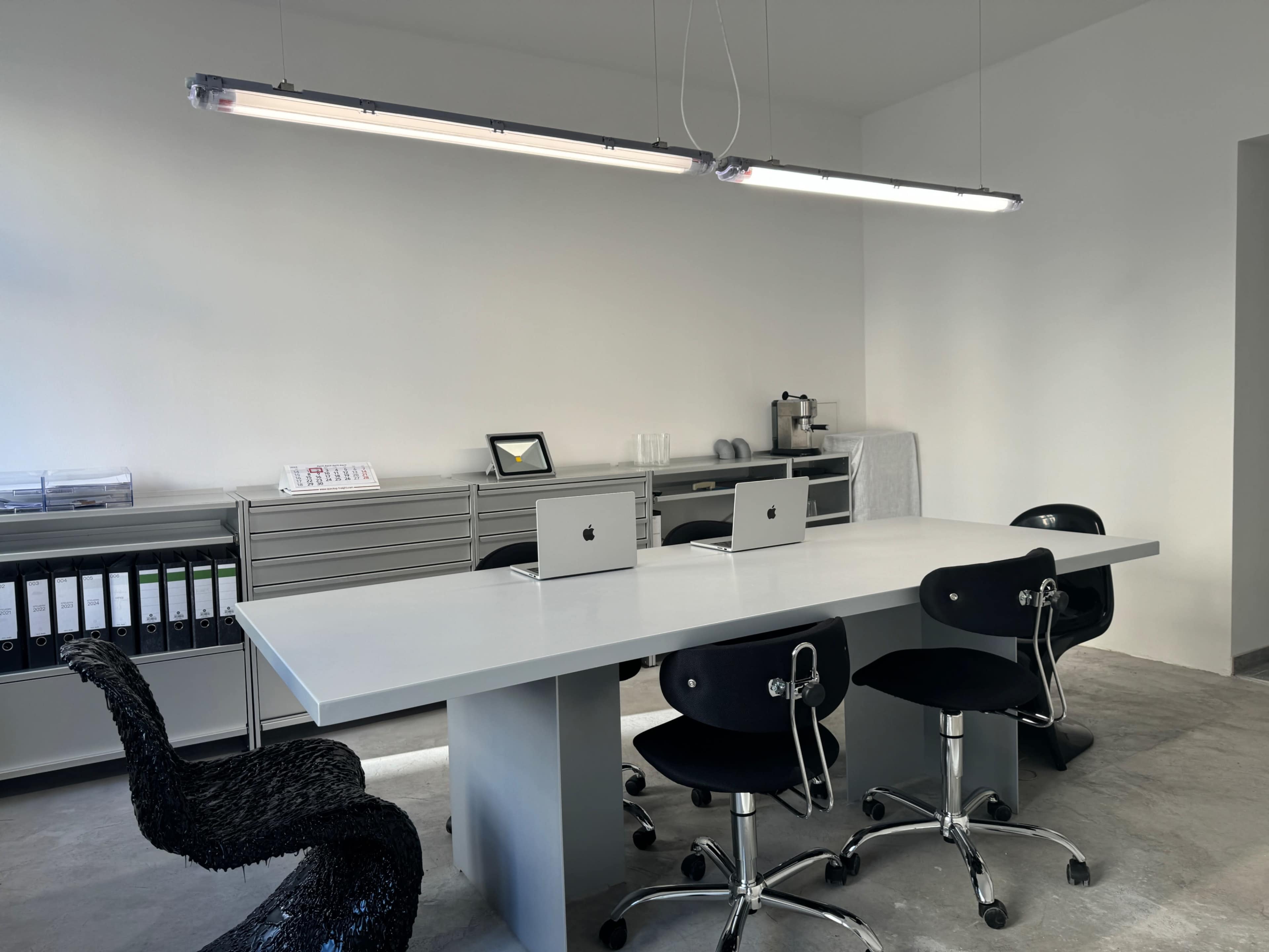 The image shows a modern office space featuring a long table with four black chairs and three Apple computers, along with storage cabinets and a coffee station in the background.