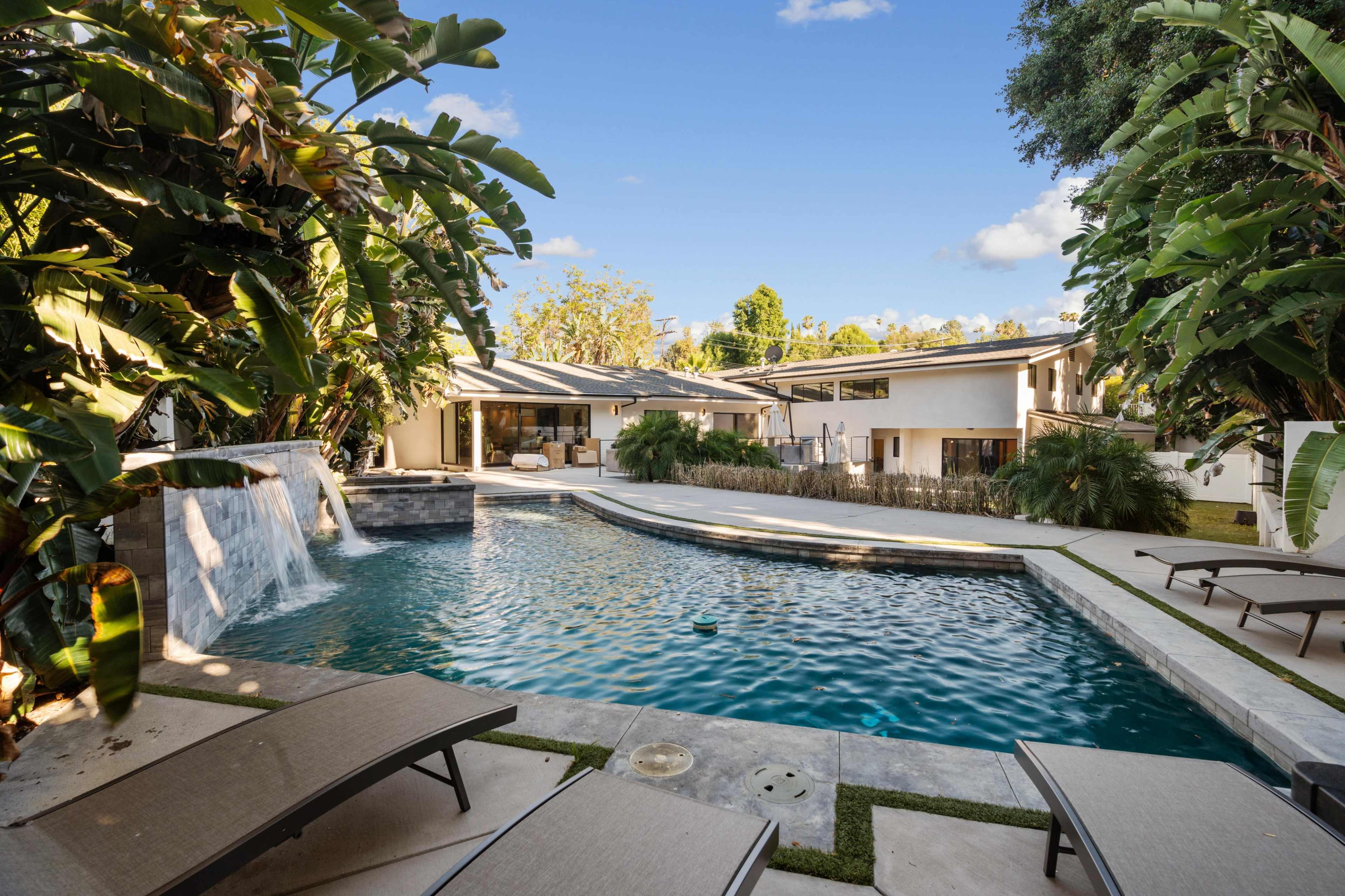 A modern backyard with a swimming pool, a waterfall feature, and lounge chairs surrounded by tropical plants and a landscaped area.