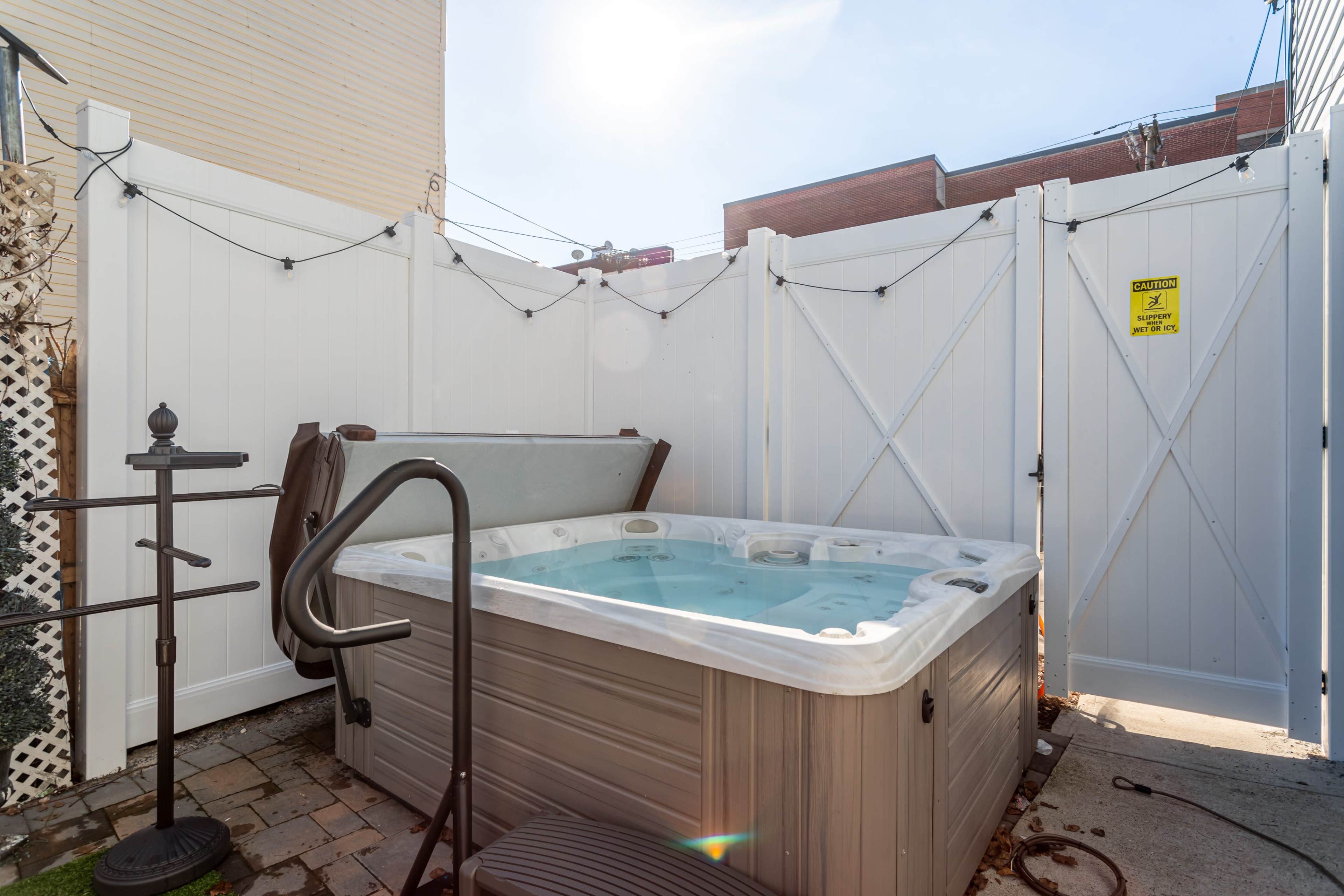 A hot tub is positioned in a small, enclosed outdoor space surrounded by white fencing and a paved floor.
