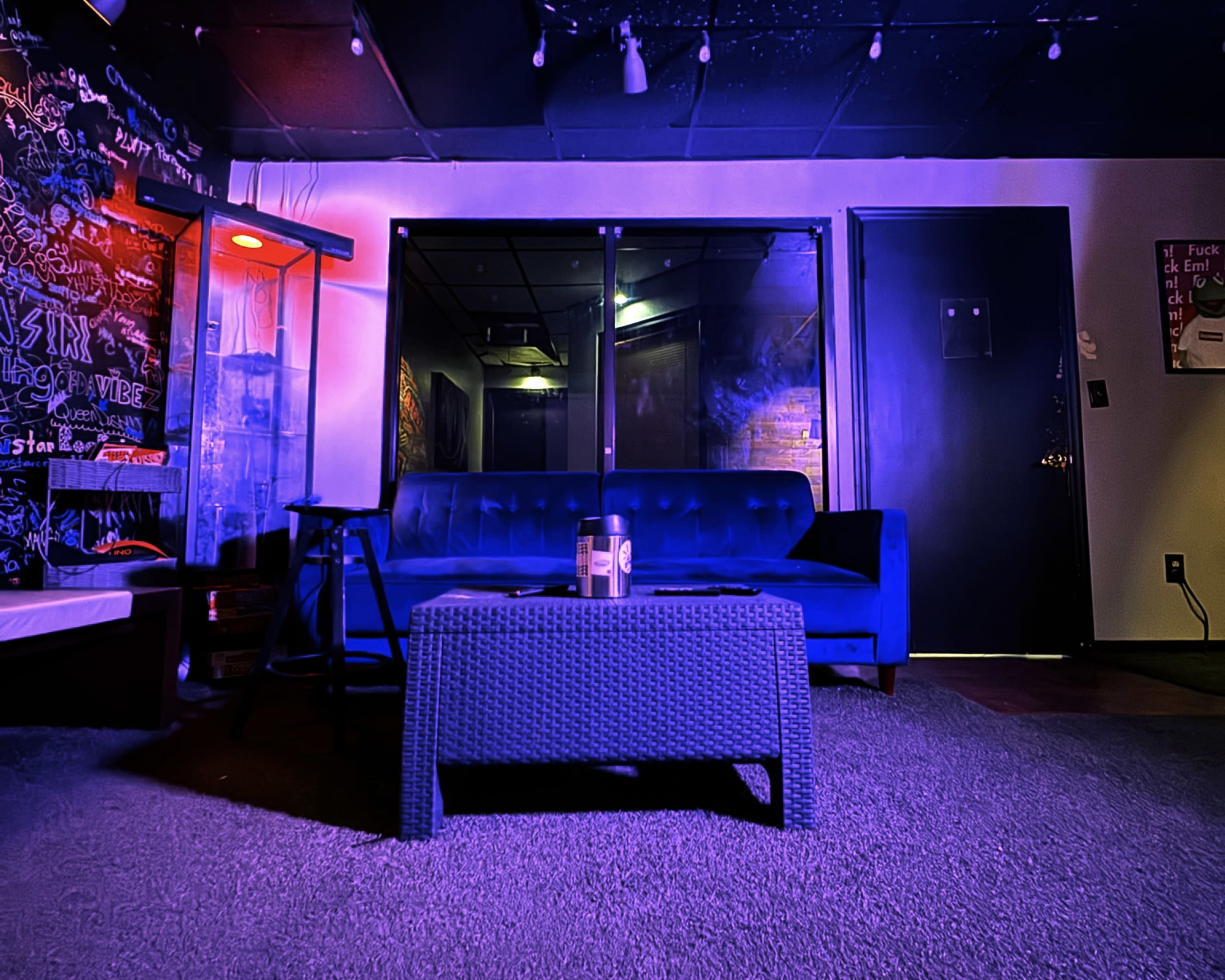 The image shows a blue couch facing a glass wall with a coffee table in front, set in a dimly lit room with graffiti-covered walls.
