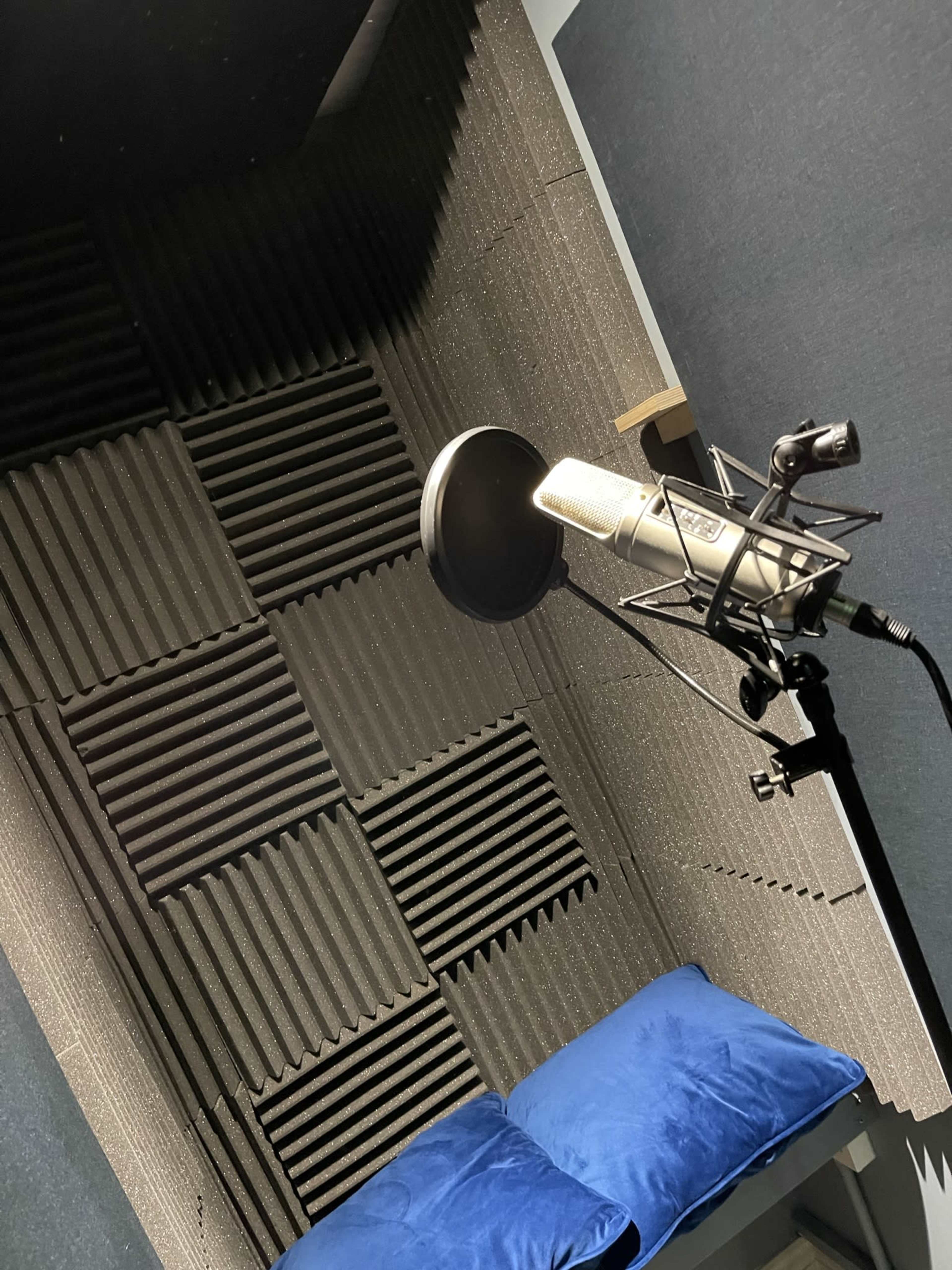 A recording studio booth featuring a microphone on a stand, with acoustic paneling and a blue cushion.