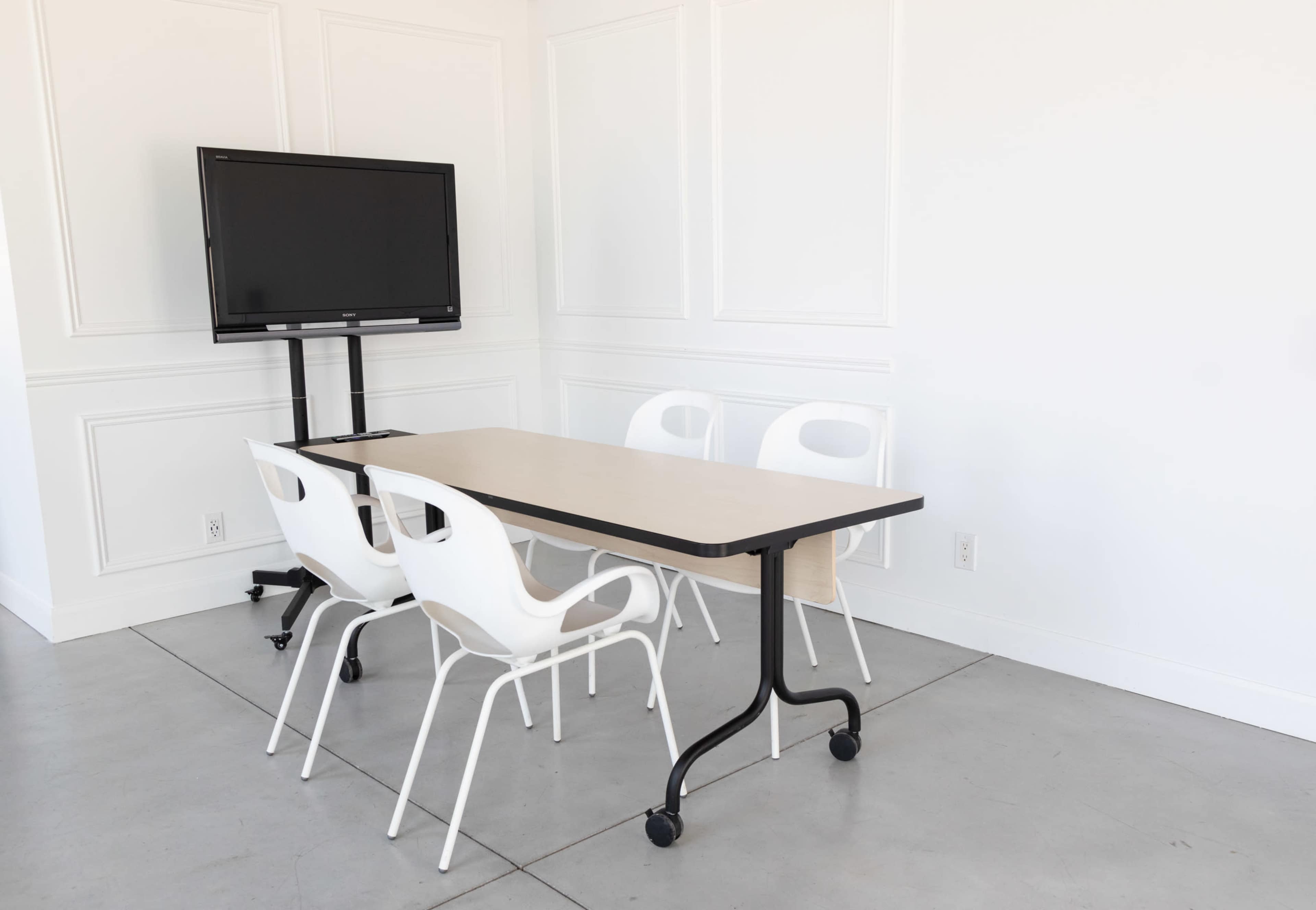 A conference room features a rectangular table surrounded by four white chairs and a mounted television on a movable stand.