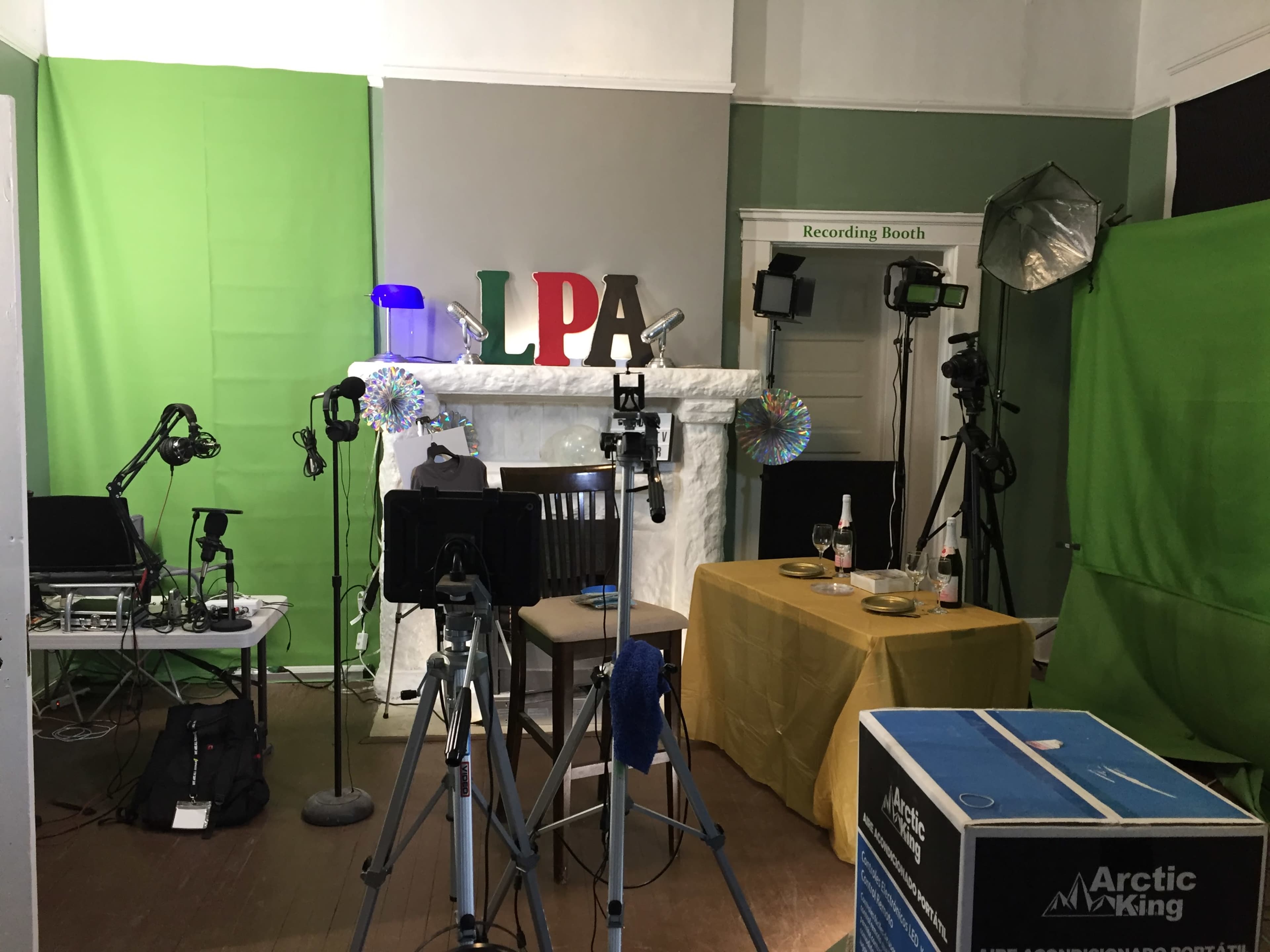 A recording studio setup with green screens, various cameras, microphones, and a table with a tablecloth, located in a room with a decorative fireplace.