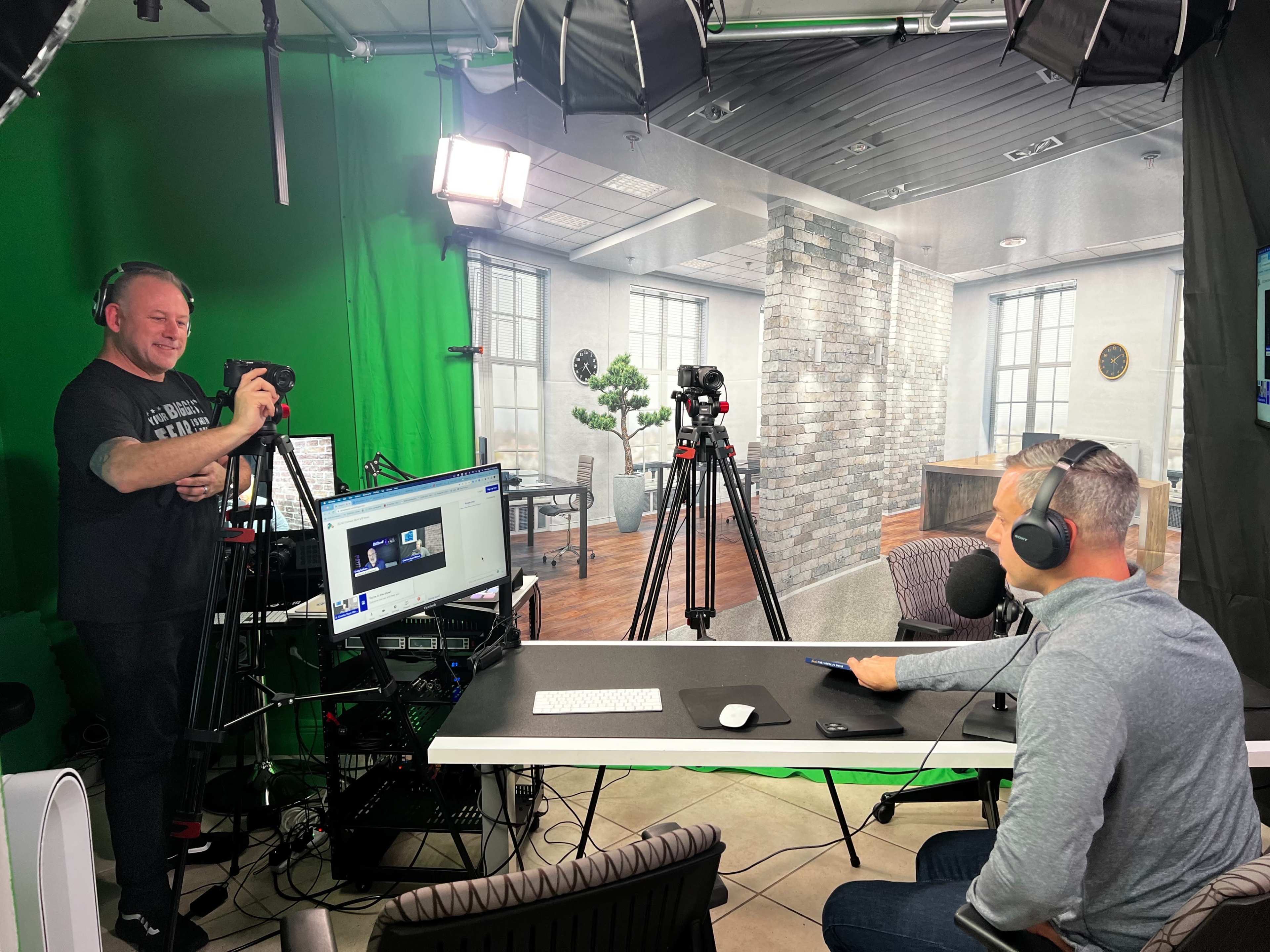 A man sits at a desk with headphones, while another man stands behind him filming with a camera in a studio setting.